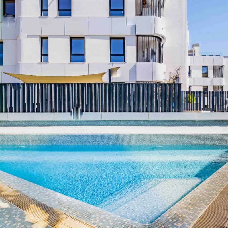 Communal swimming pool featuring blue mosaic tiling and integrated steps, set against a modern white residential building with sleek metal fencing and Mediterranean landscaping.