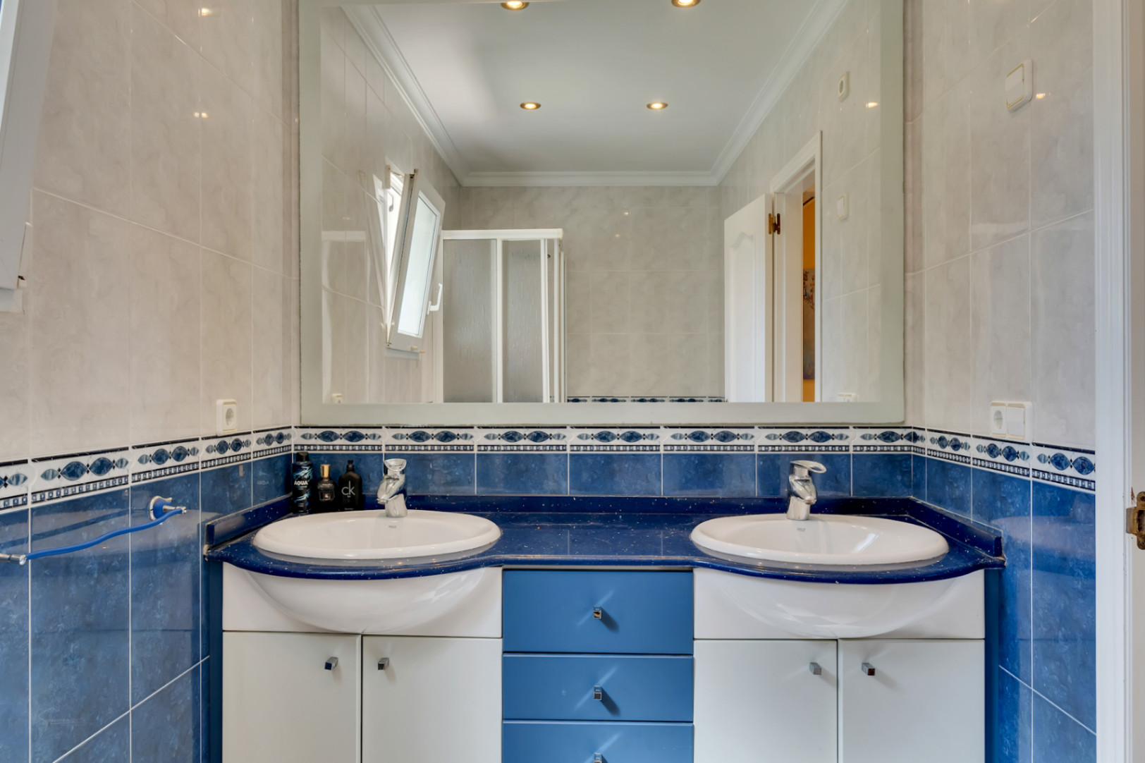 Bathroom featuring a double vanity with a blue countertop, integrated white sinks, and chrome fixtures. The space includes blue wall tiles with decorative borders and a large mirror.