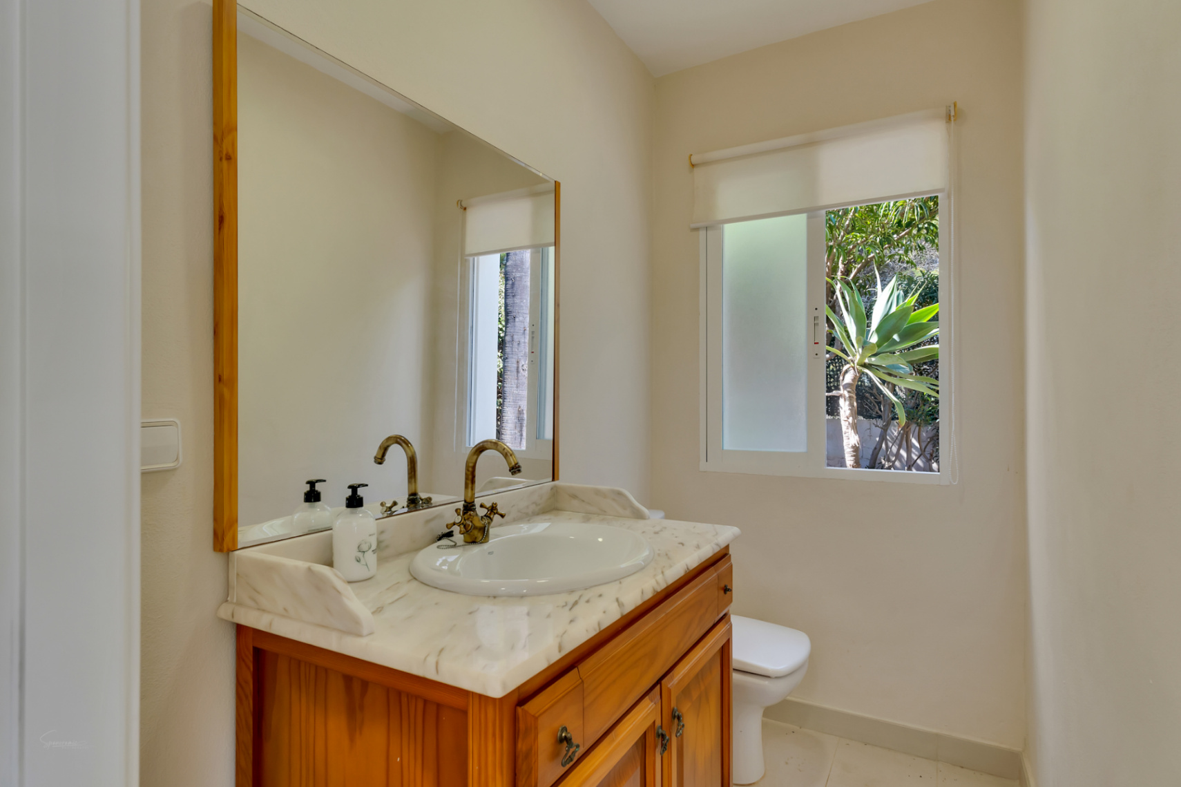 Bathroom featuring a wooden vanity with marble countertop and vintage-style brass faucet. A window provides natural light and views of the garden greenery.
