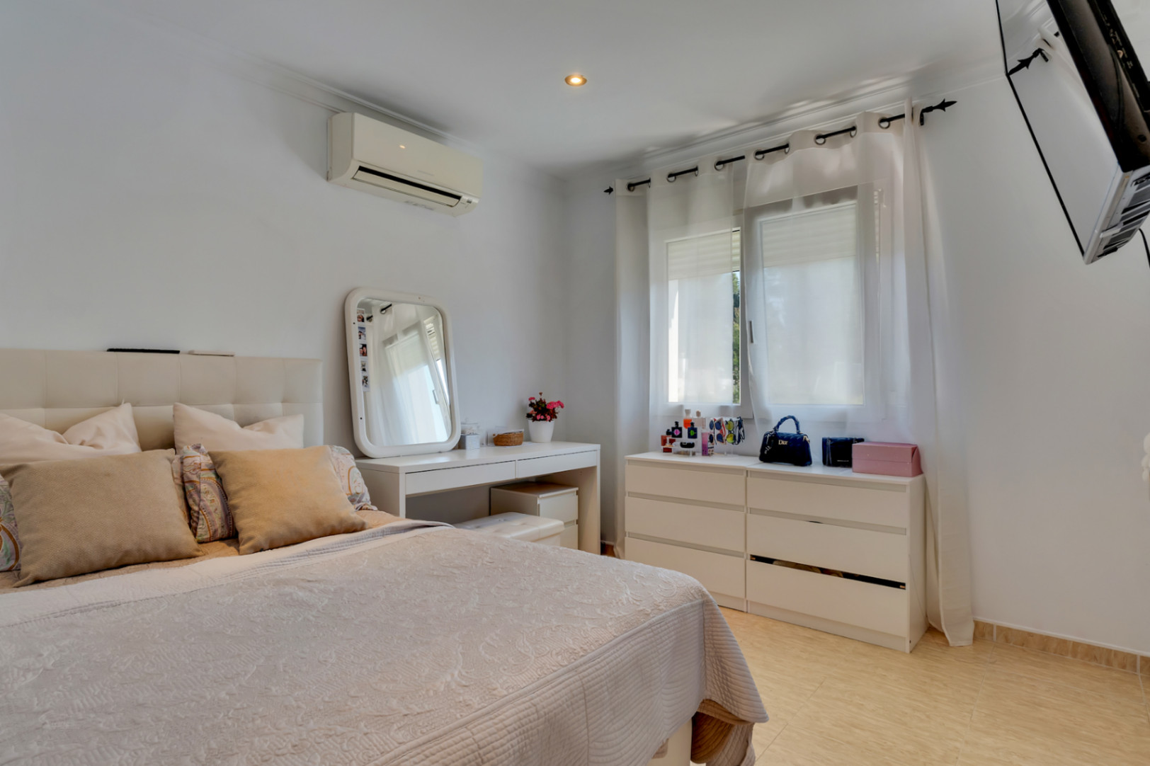 Bright bedroom featuring a tufted headboard, white dressing table, and matching drawers. Equipped with split air conditioning, tiled flooring, and a wall-mounted TV.
