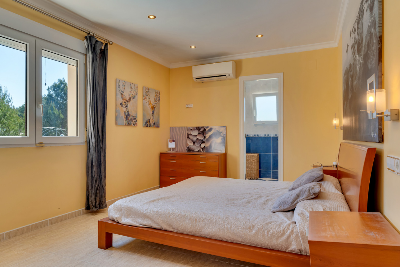 Bright bedroom featuring split-system air conditioning, tiled flooring, and an ensuite bathroom. Large PVC windows provide natural light and views of the surrounding greenery.