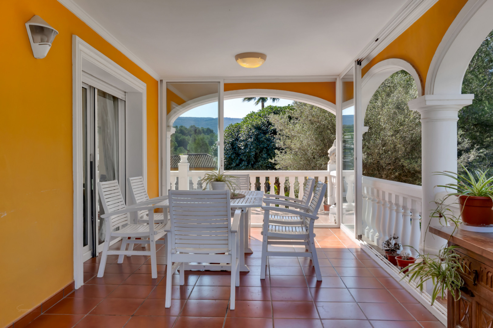 Covered terrace featuring a white wooden dining set, terracotta tile flooring, and glass-enclosed arches overlooking the lush Mediterranean landscape.