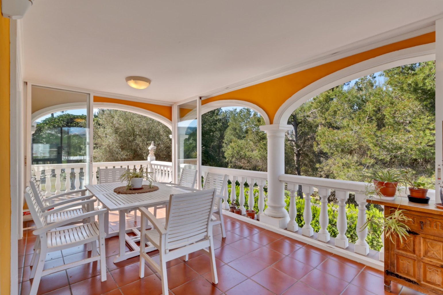 Covered terrace featuring arched openings, terracotta flooring, and glass enclosures. Includes a white wooden dining set overlooking the lush pine forest.