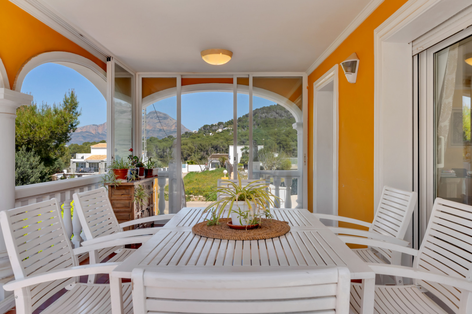 Covered terrace featuring glass curtains, a white wooden dining set, and open views of the surrounding mountains and Mediterranean landscape.