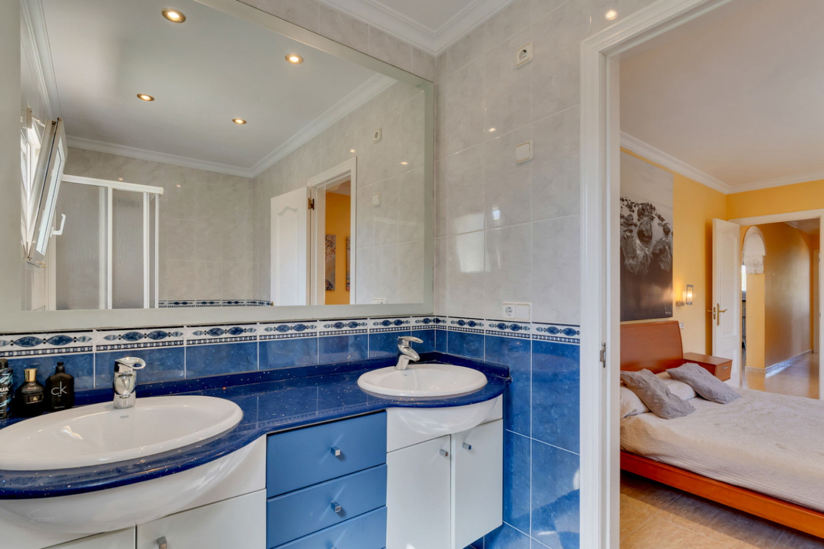 En-suite bathroom featuring a double vanity with a blue countertop, matching wall tiles, and a large mirror with recessed lighting, connected to a bedroom.