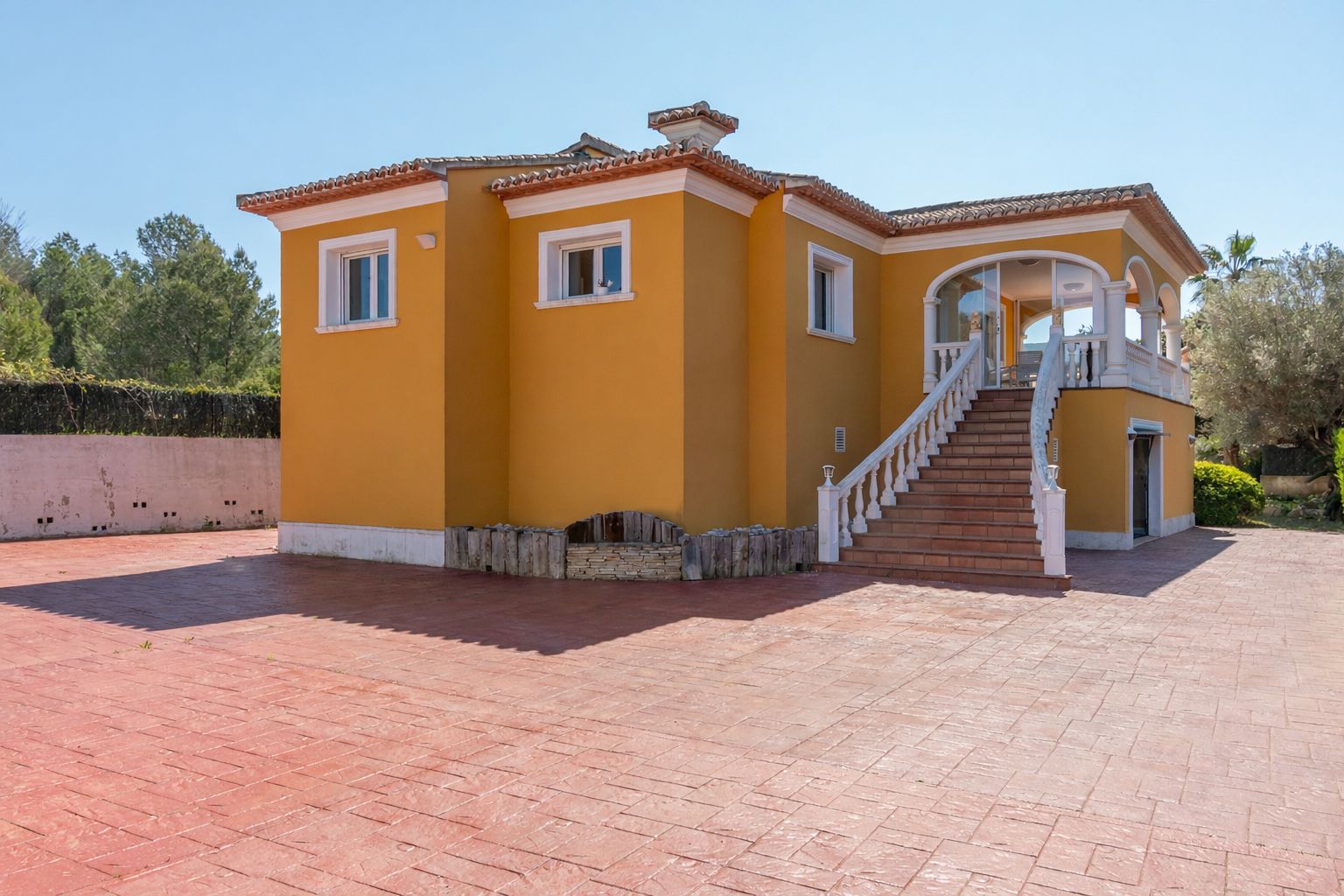 Exterior view of the ochre-toned villa featuring a spacious paved driveway and a grand staircase with white balustrades leading to the elevated arched terrace.