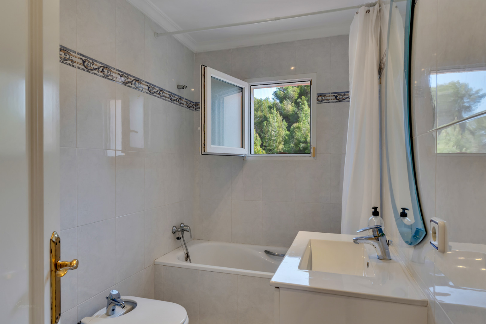 Functional bathroom featuring a bathtub, white vanity unit, and bidet. A tilt-and-turn window provides natural light and views of the surrounding Mediterranean greenery.