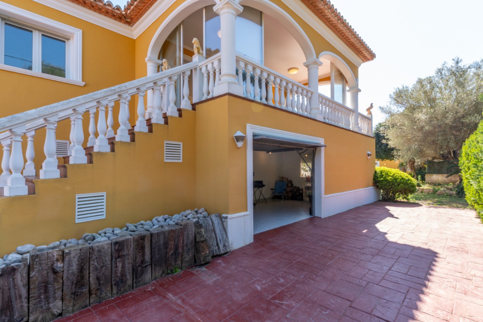 Mediterranean-style facade featuring an external staircase with white balustrades leading to an arched terrace above an integrated garage and paved courtyard.
