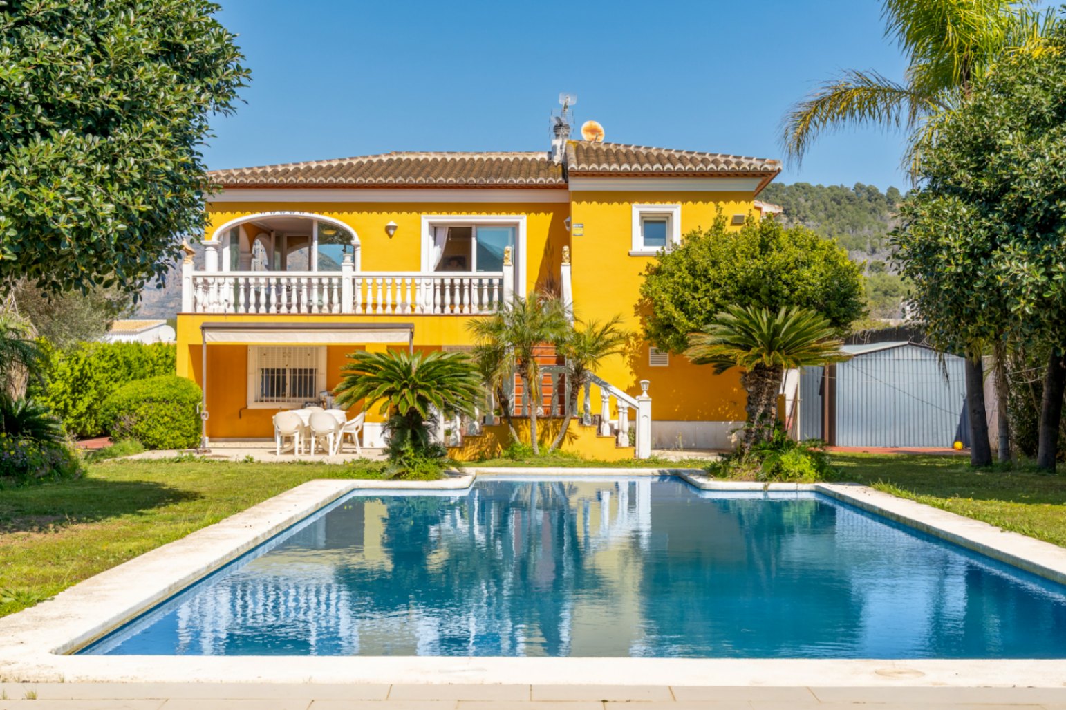 Mediterranean villa featuring a yellow facade, a large swimming pool, and an upper terrace with white balustrades overlooking a landscaped garden.