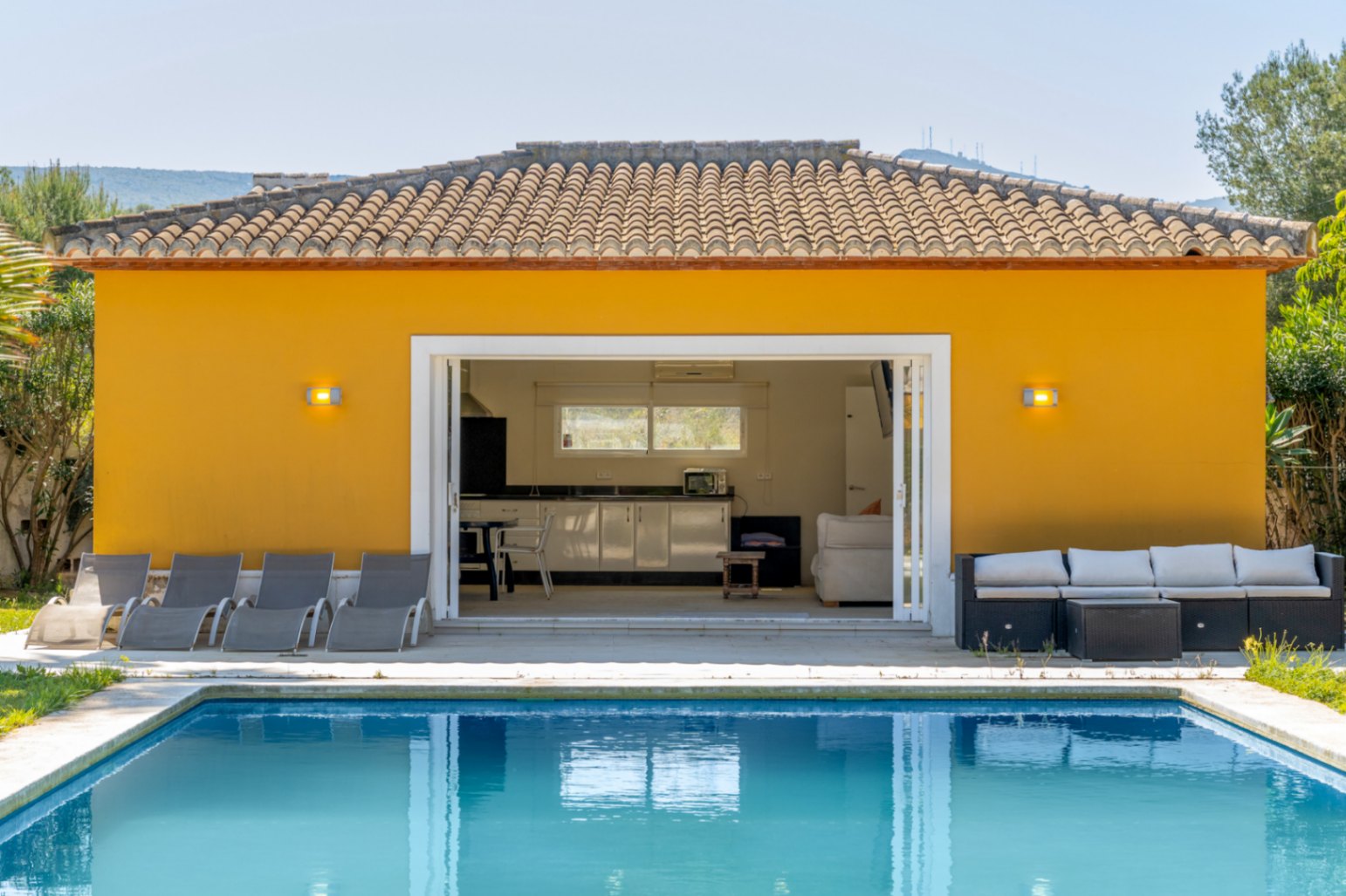 Poolside annex featuring a fully equipped kitchen, living area, and large sliding doors opening onto the sun terrace with loungers and outdoor furniture.