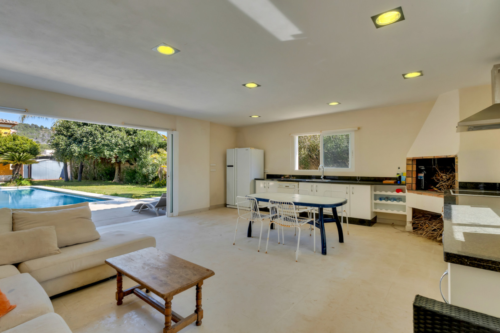 Spacious open-plan summer kitchen and living area featuring a built-in barbecue, with direct access to the swimming pool and garden through sliding doors.