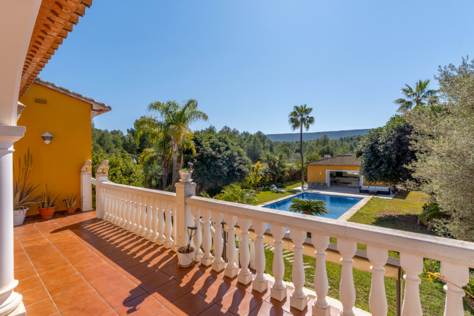 Upper terrace with terracotta tiling and white balustrades, overlooking the large swimming pool, landscaped gardens, and the surrounding lush green nature of Jávea.