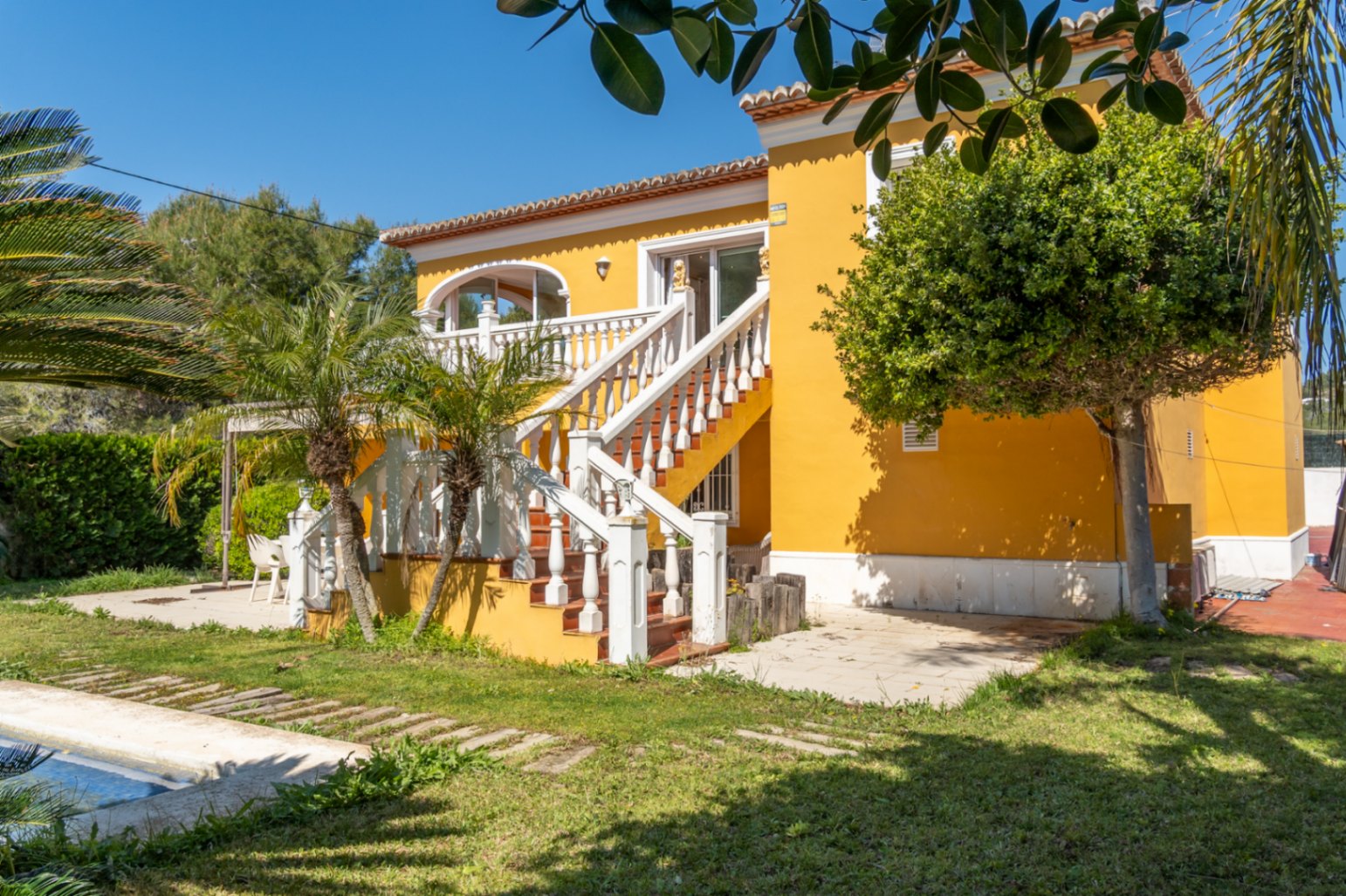 Yellow villa exterior featuring a grand staircase with white balustrades leading to an upper terrace. The garden includes native palms and a swimming pool edge.