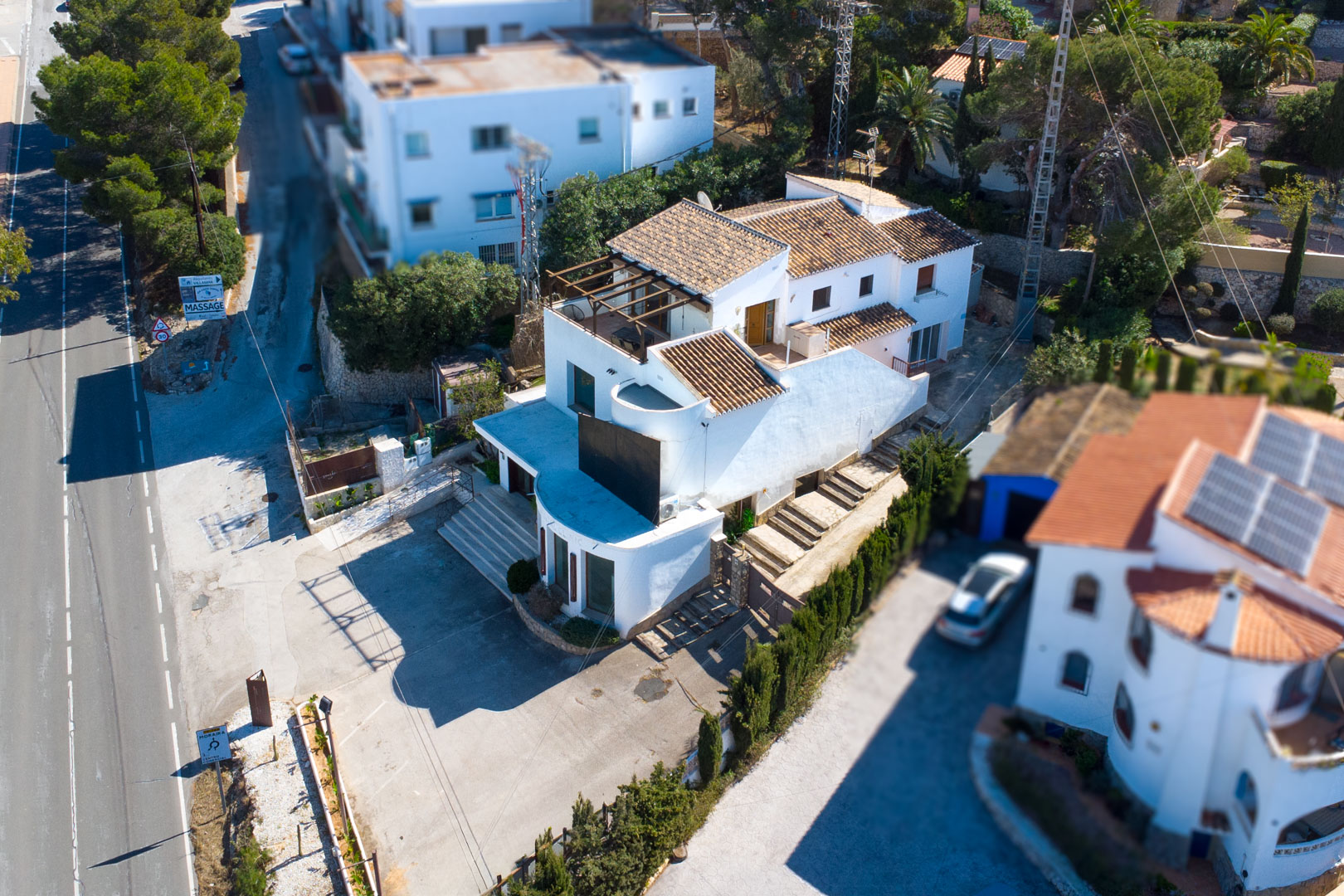 Aerial view of the multi-level property featuring white facades and terracotta roofs. The image highlights the spacious front parking area and its prominent location by the road.