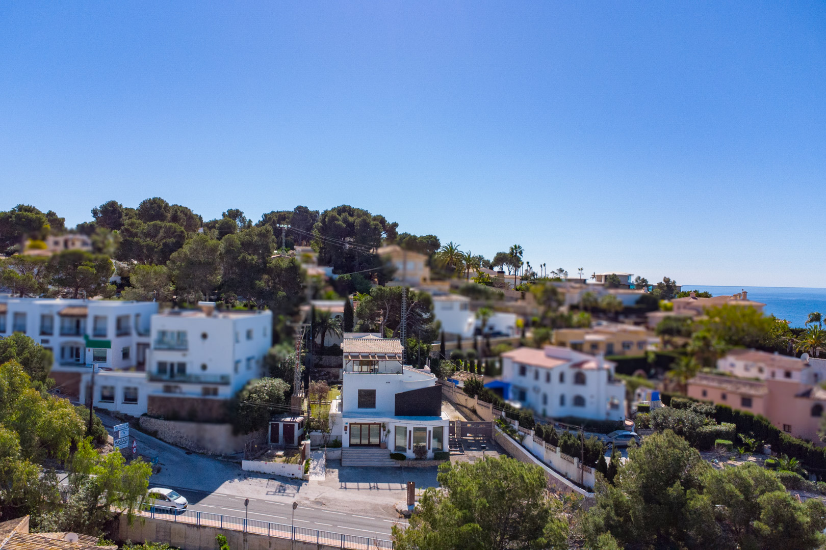 Aerial view of the three-story property located roadside, featuring a white facade, large commercial windows, and a top-floor terrace with a wooden pergola.