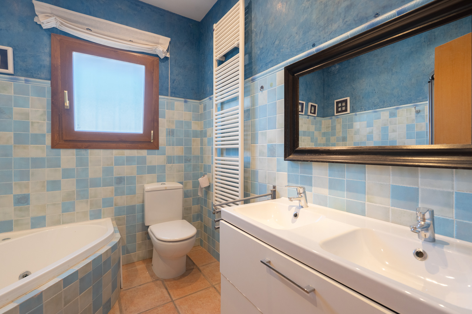 Bathroom featuring blue-toned tiling, a double vanity, corner bathtub, and a vertical towel radiator against textured blue walls.