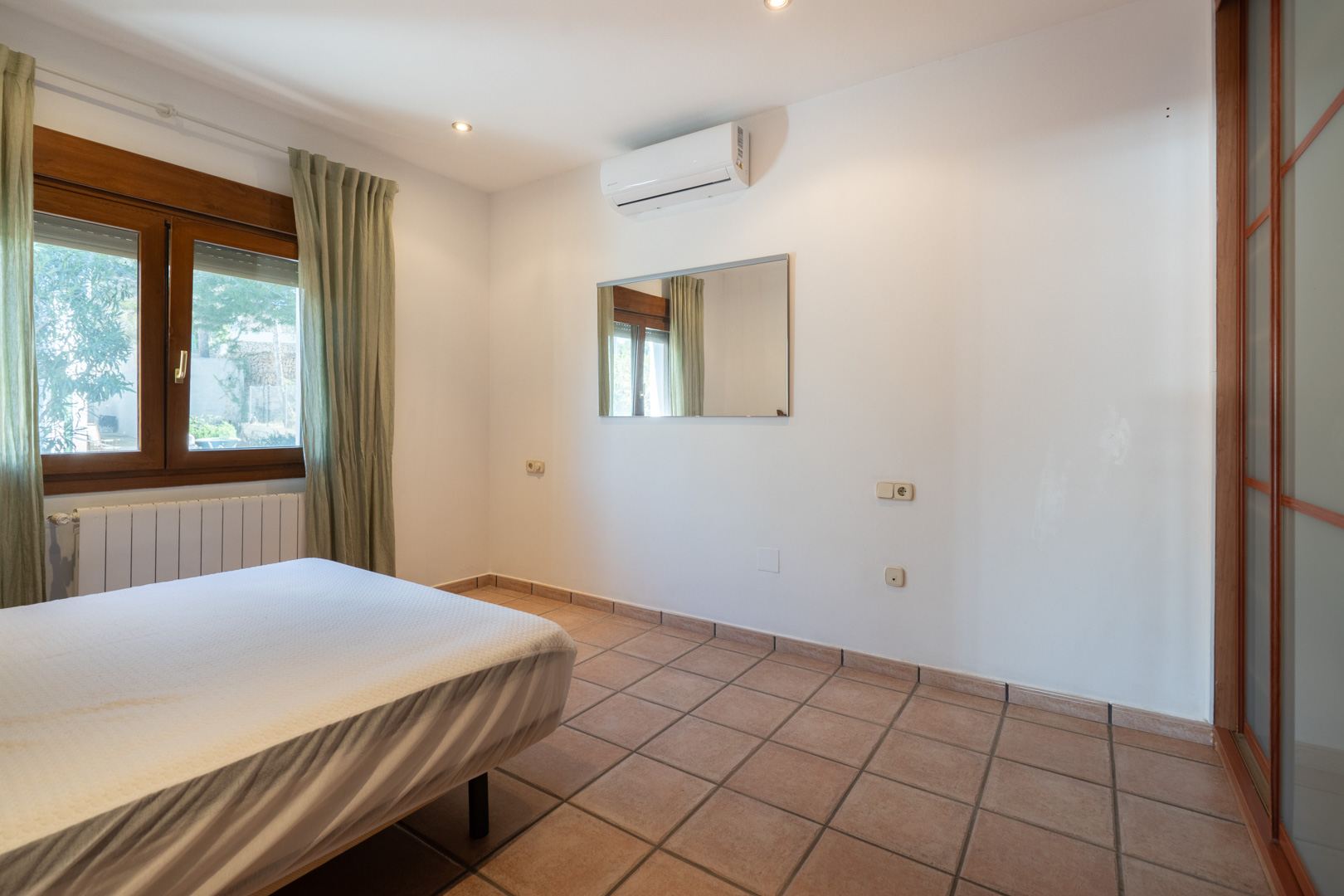 Bedroom featuring terracotta-style floor tiles, split air conditioning, and a wooden window frame. Includes recessed lighting and a sliding door closet.