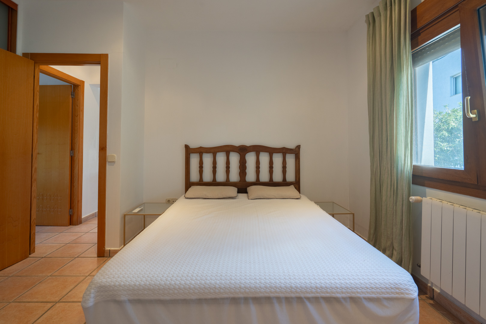 Bedroom featuring terracotta tile flooring, a classic wooden headboard, and natural light from a wood-framed window. Minimalist glass side tables and a radiator are included.