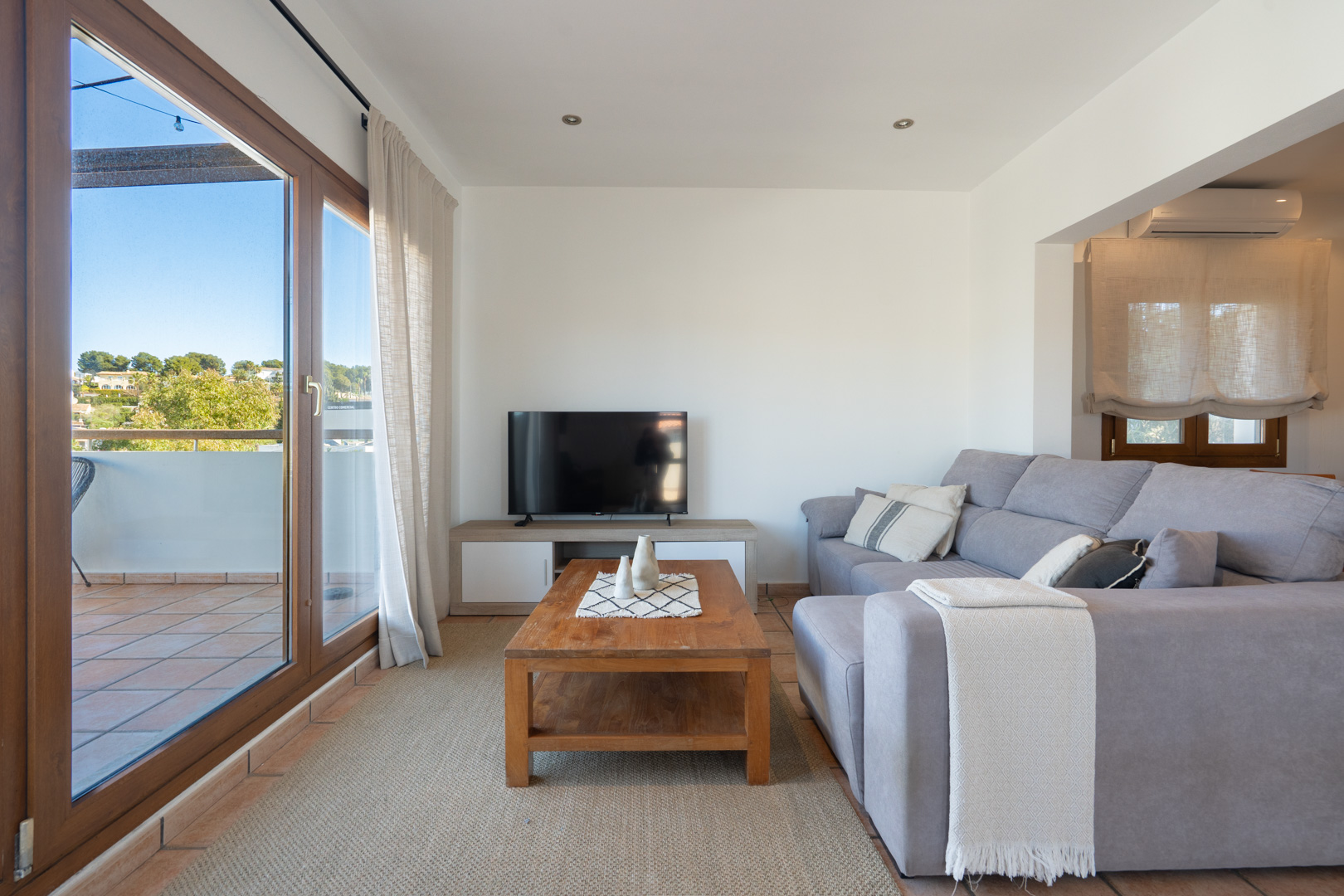 Bright living room featuring a grey sectional sofa, wooden coffee table, and large sliding glass doors opening onto a private terrace with scenic views.