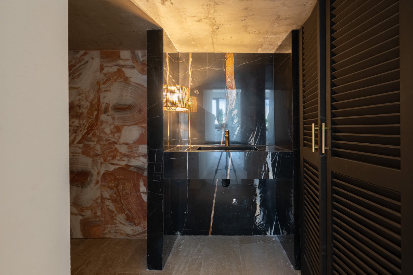 Designer washroom featuring an integrated black marble sink, gold fixtures, and natural stone wall cladding. Includes warm pendant lighting and dark louvered doors.