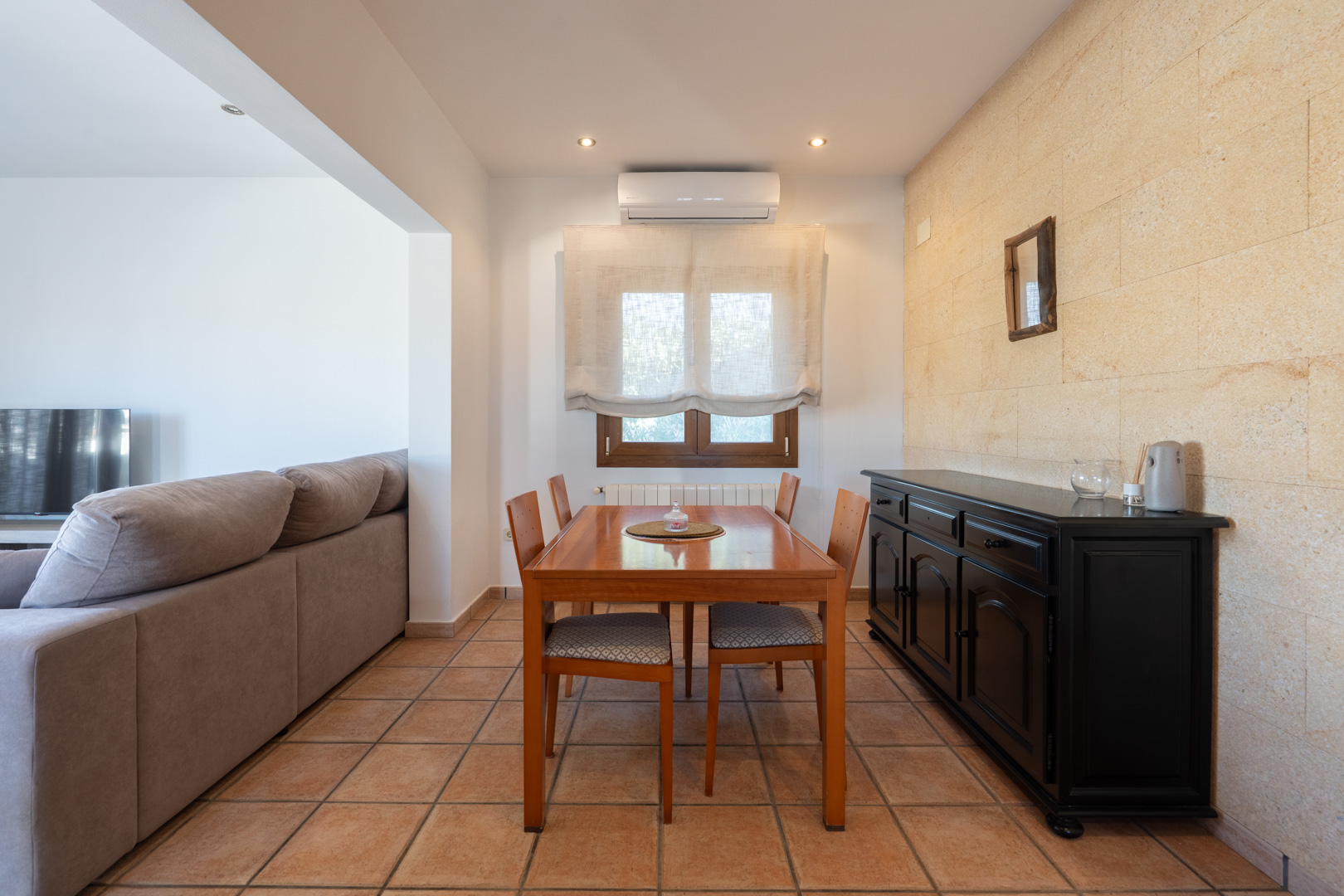 Dining area featuring a natural stone accent wall, rustic tile flooring, a wooden table for four, and a black sideboard. Equipped with air conditioning and a radiator.