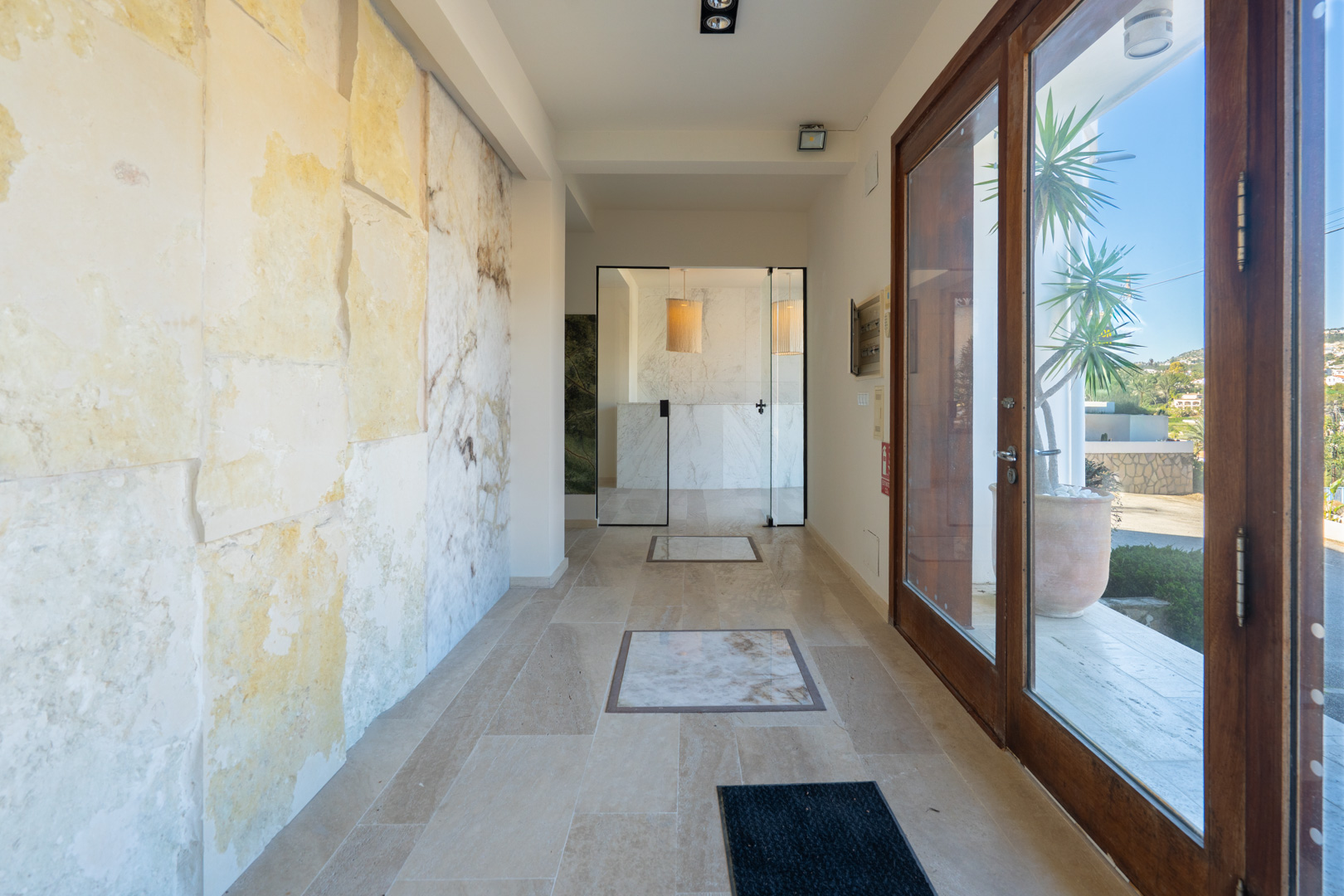 Entrance hall featuring natural stone wall panels, travertine flooring with marble inlays, and a glass partition leading to the reception area.