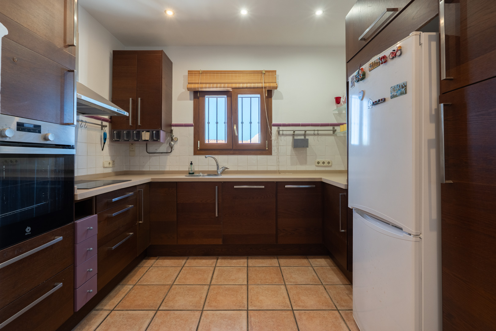 Equipped kitchen with dark wood cabinetry, cream countertops, and ceramic tile flooring. Features integrated appliances and a window providing natural light.