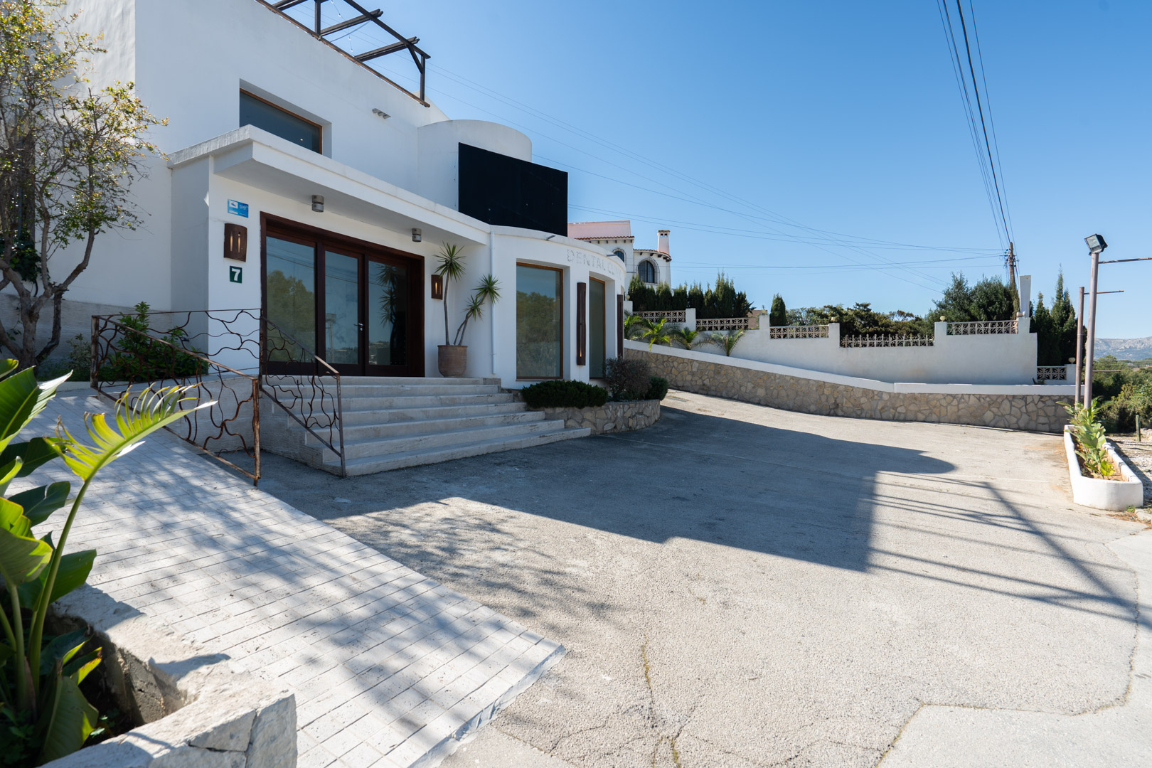 Exterior facade featuring a glazed main entrance, wide stone steps, an accessibility ramp, and a paved parking area under a clear blue sky.