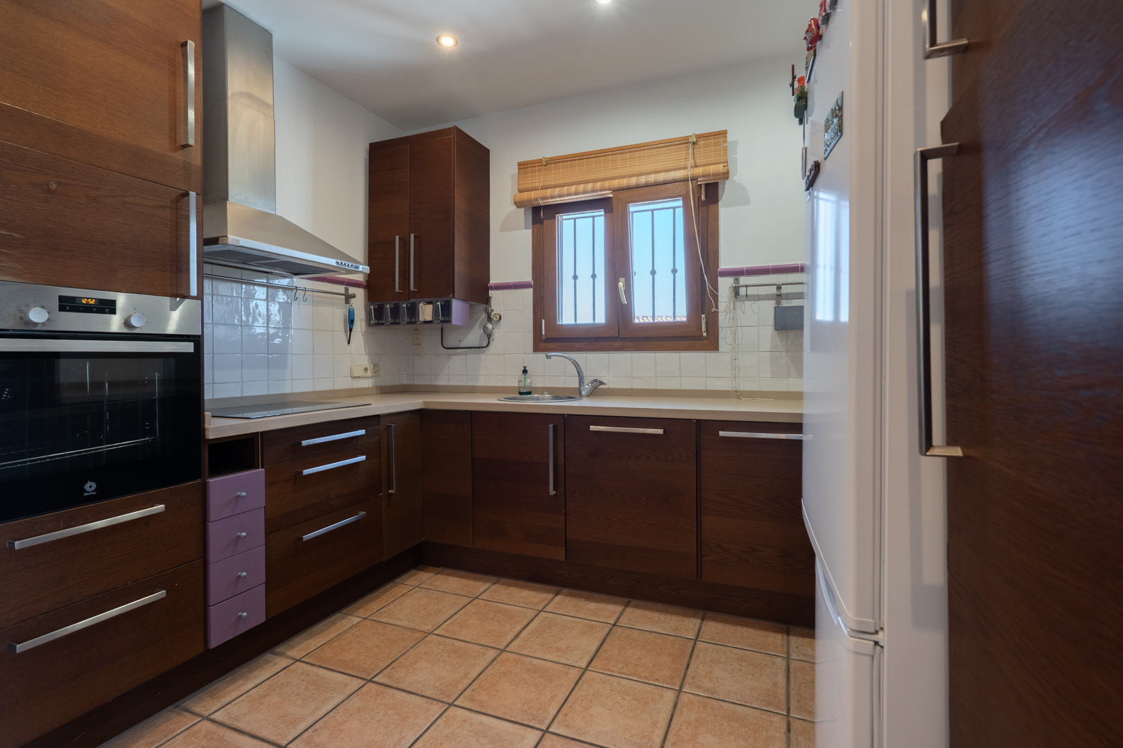 Functional kitchen featuring dark wood cabinetry, integrated appliances, and terracotta floor tiles. A window above the sink provides natural light.