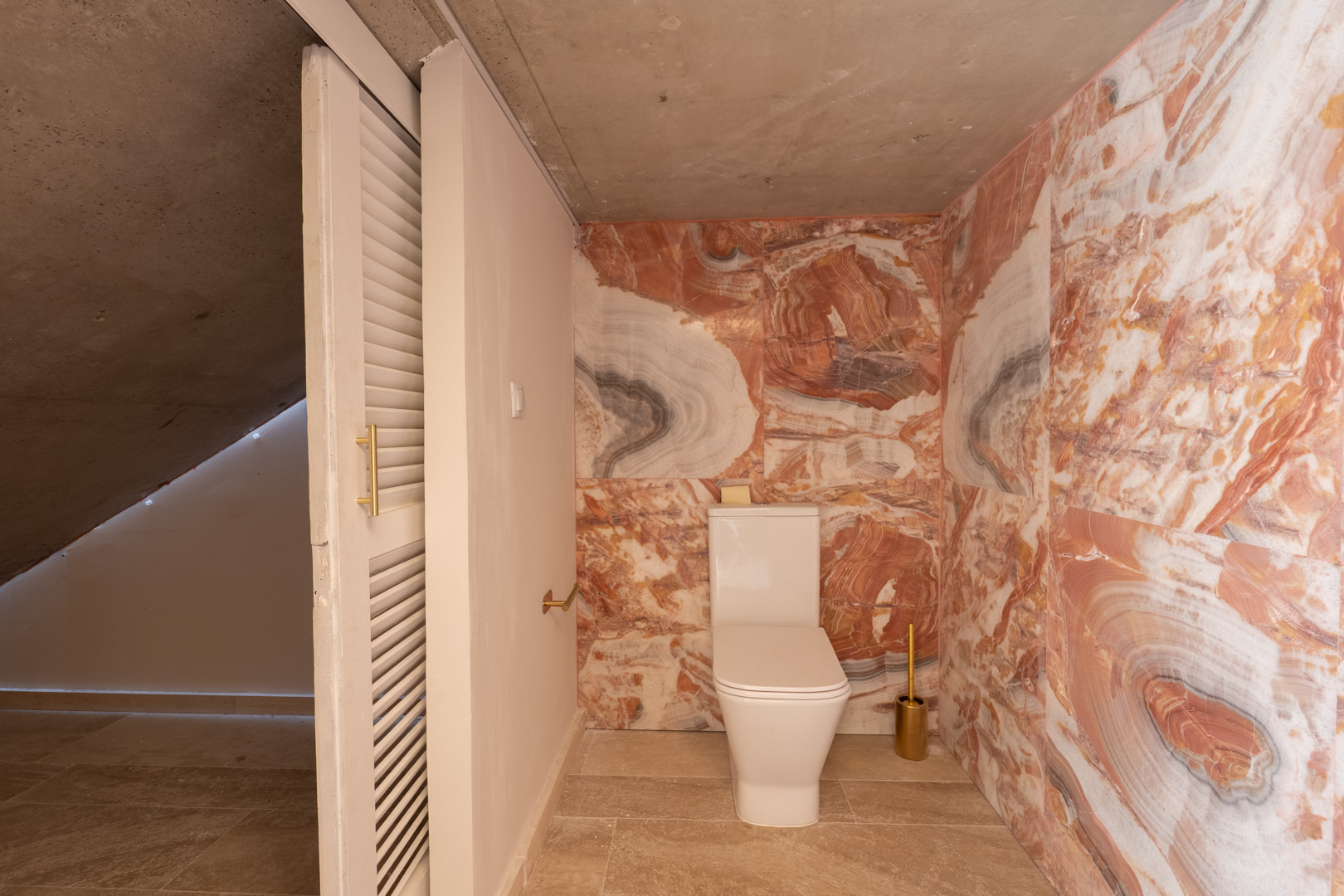 Guest toilet featuring striking red-veined marble walls, a modern white toilet, and exposed concrete ceiling, complemented by gold-toned accessories.