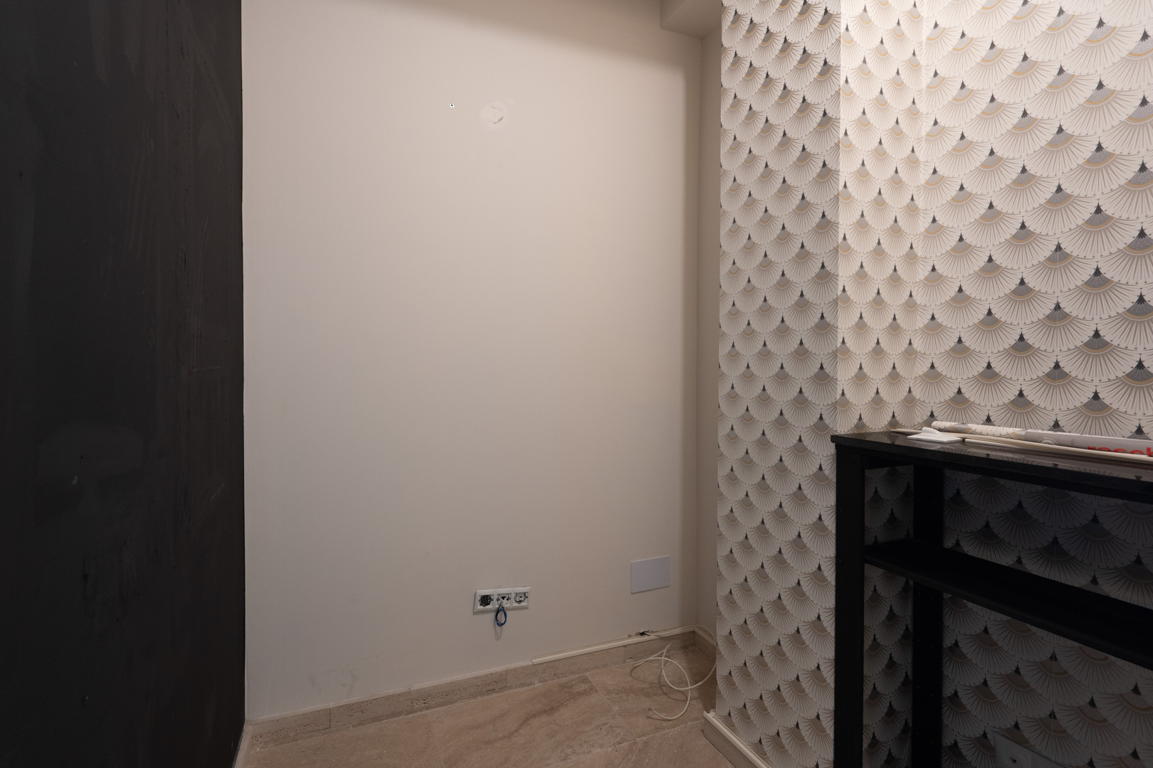 Interior corner featuring Art Deco patterned wallpaper, neutral off-white walls with electrical outlets, and light-toned stone flooring.