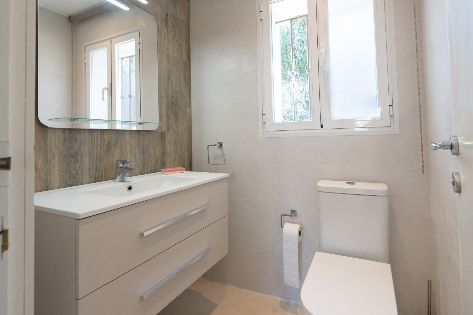 Modern bathroom featuring a floating vanity with dual drawers, wood-effect accent wall, large mirror with glass shelf, and a white PVC window.