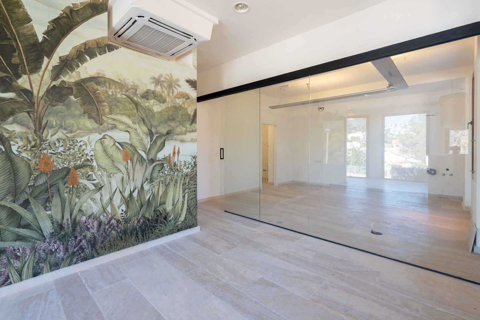 Open-plan commercial space featuring stone tile flooring, a tropical wall mural, glass partitions with black frames, and ceiling-mounted air conditioning.