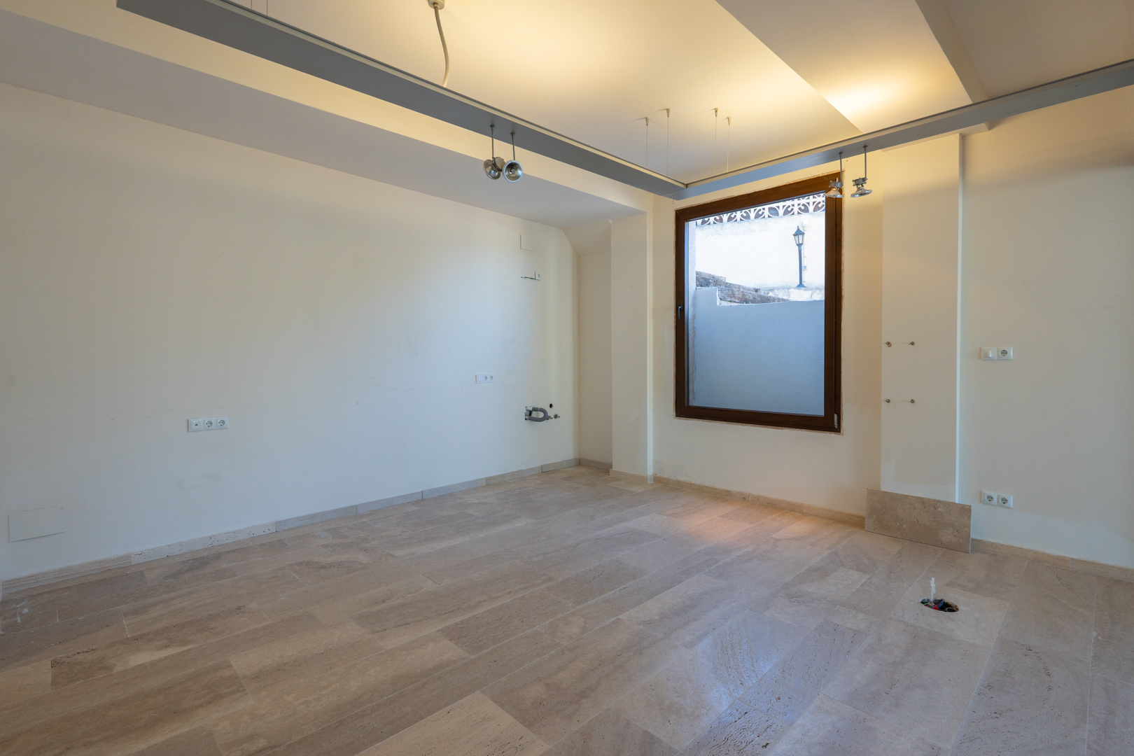 Open-plan interior featuring natural stone flooring, recessed ceiling lighting with tracks, and a large wood-framed window. Includes wall-mounted electrical and plumbing connections.