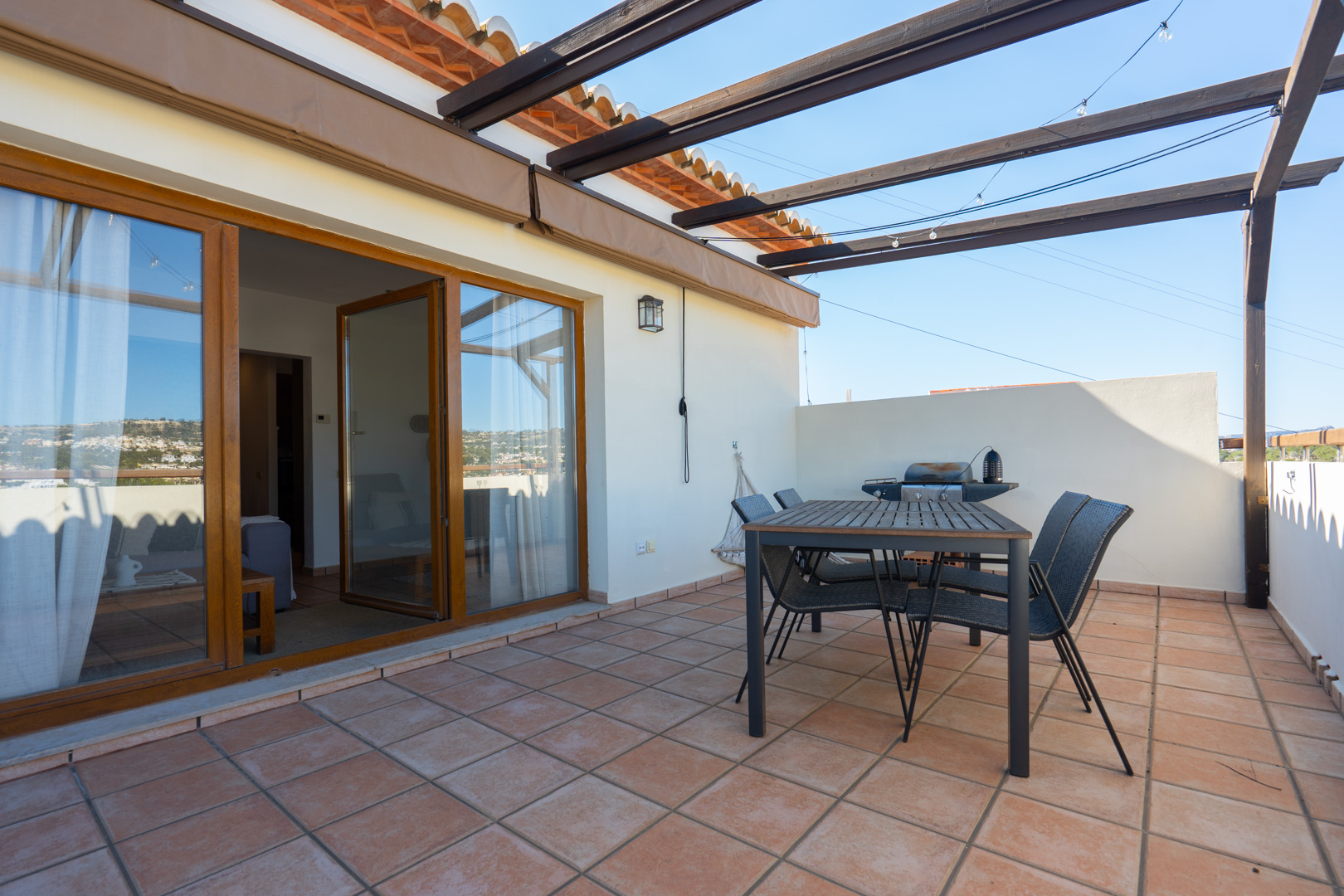 Outdoor terrace featuring terracotta tiling, a wooden dining set under a pergola, and a barbecue area. Large glass doors with wooden frames connect to the interior living space.