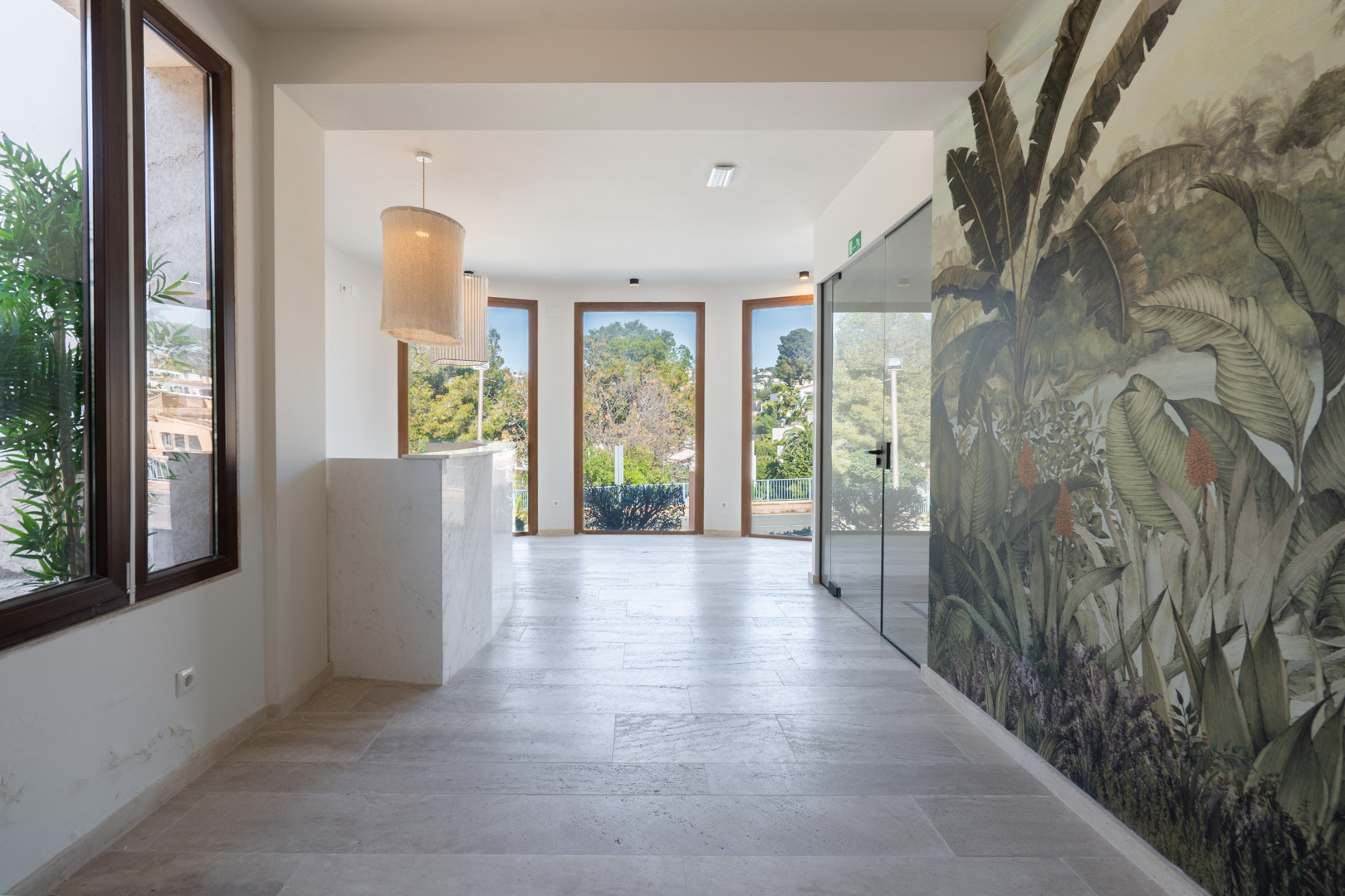 Reception hall featuring a white marble desk, natural stone flooring, and a tropical mural. Large wood-framed windows provide abundant natural light.
