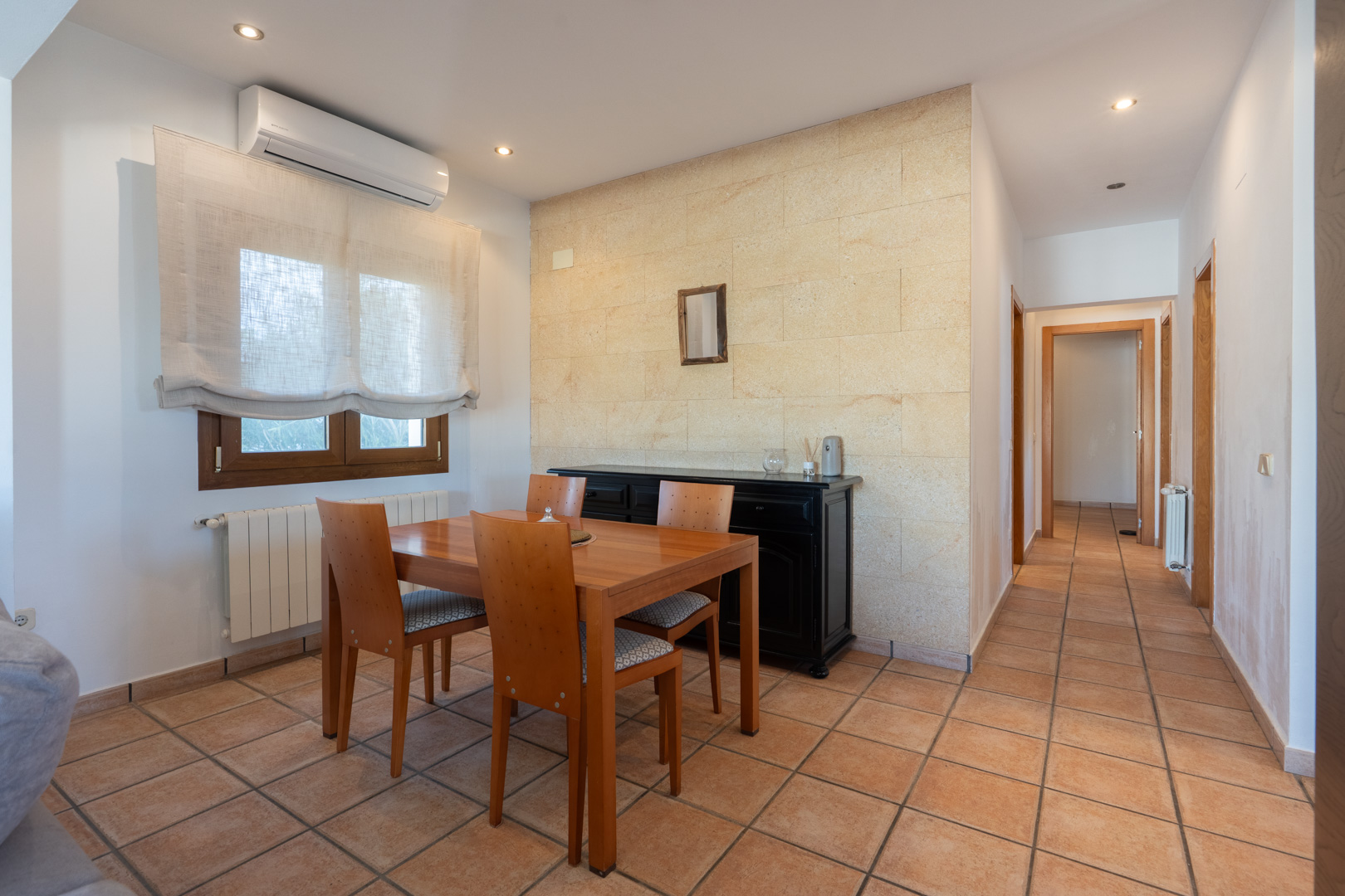 Residential dining area featuring a natural stone accent wall, terracotta tile flooring, air conditioning, and a hallway leading to bedrooms.