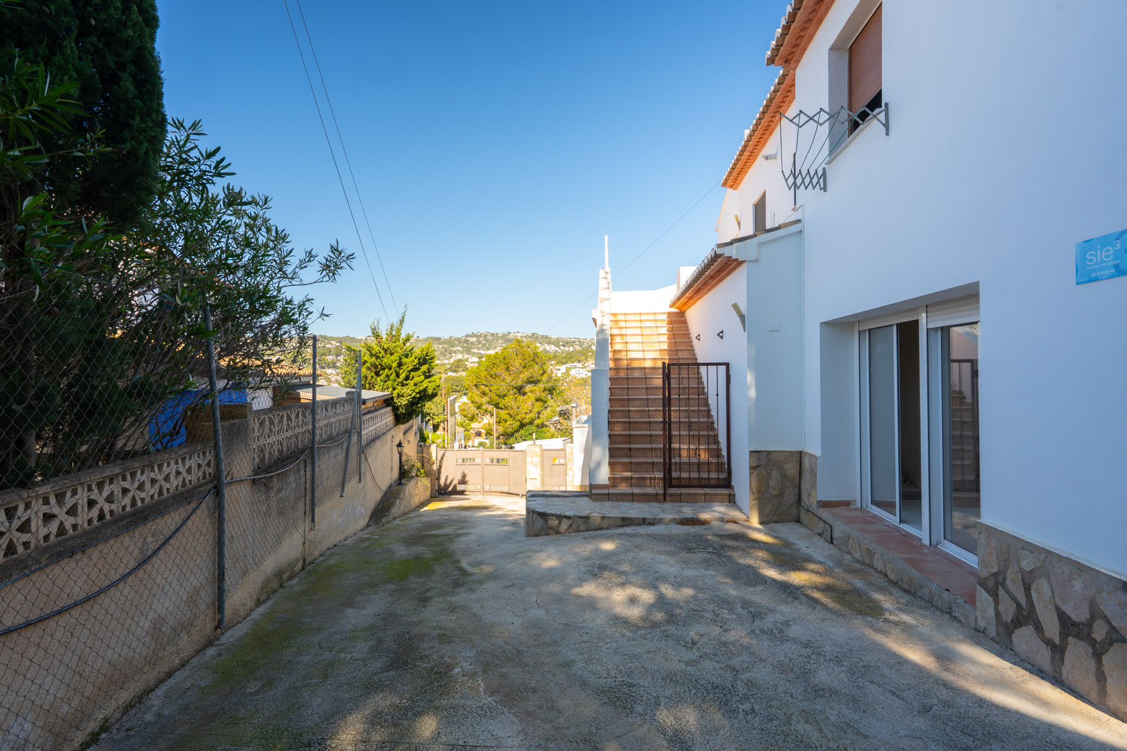 Side entrance featuring a paved parking area, external tiled staircase for independent upper-floor access, and a white facade with natural stone cladding.
