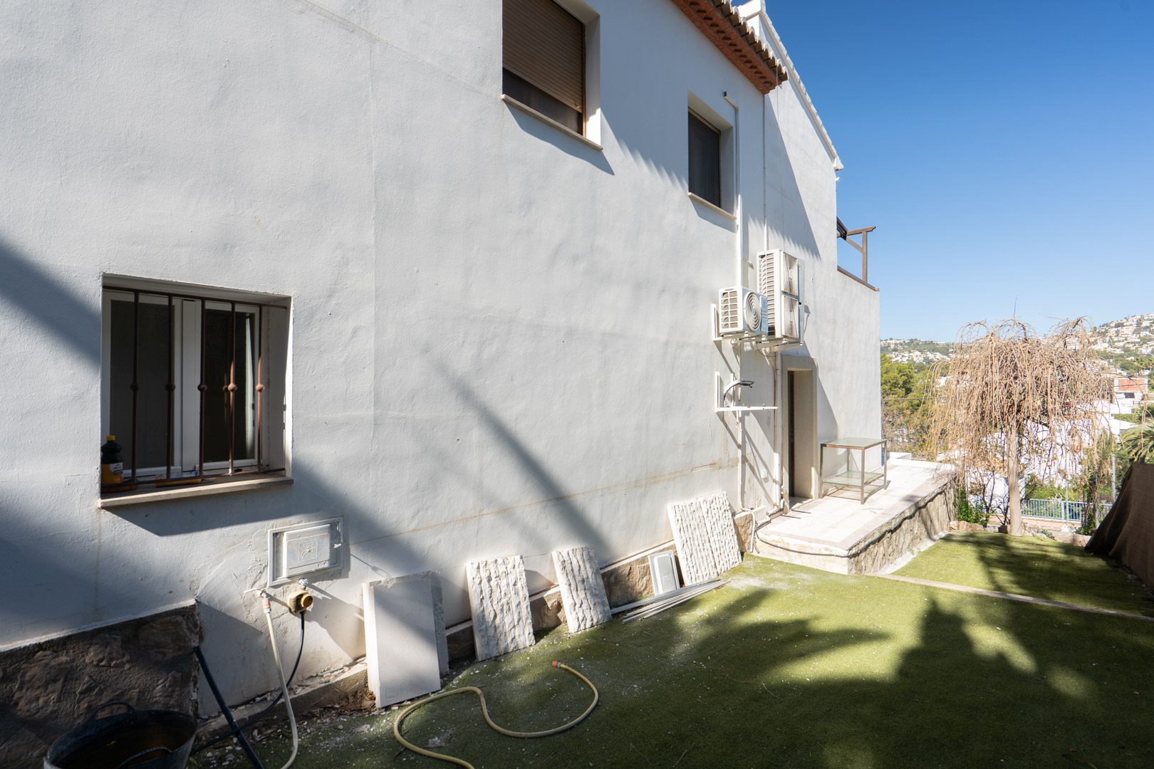 Side exterior featuring white plastered walls, natural stone base, external air conditioning units, and a garden area with artificial grass and open views.