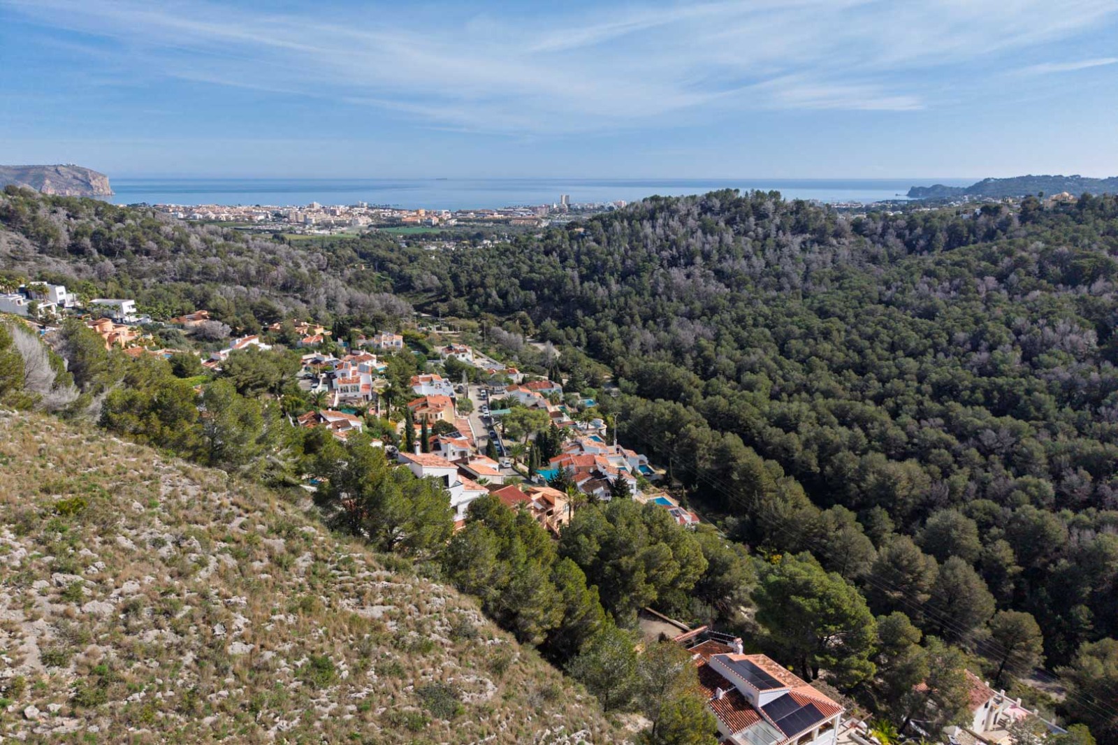 Panoramic aerial view showcasing the elevated topography over wooded hills, with clear vistas of the Mediterranean Sea and the distant Jávea coastline.
