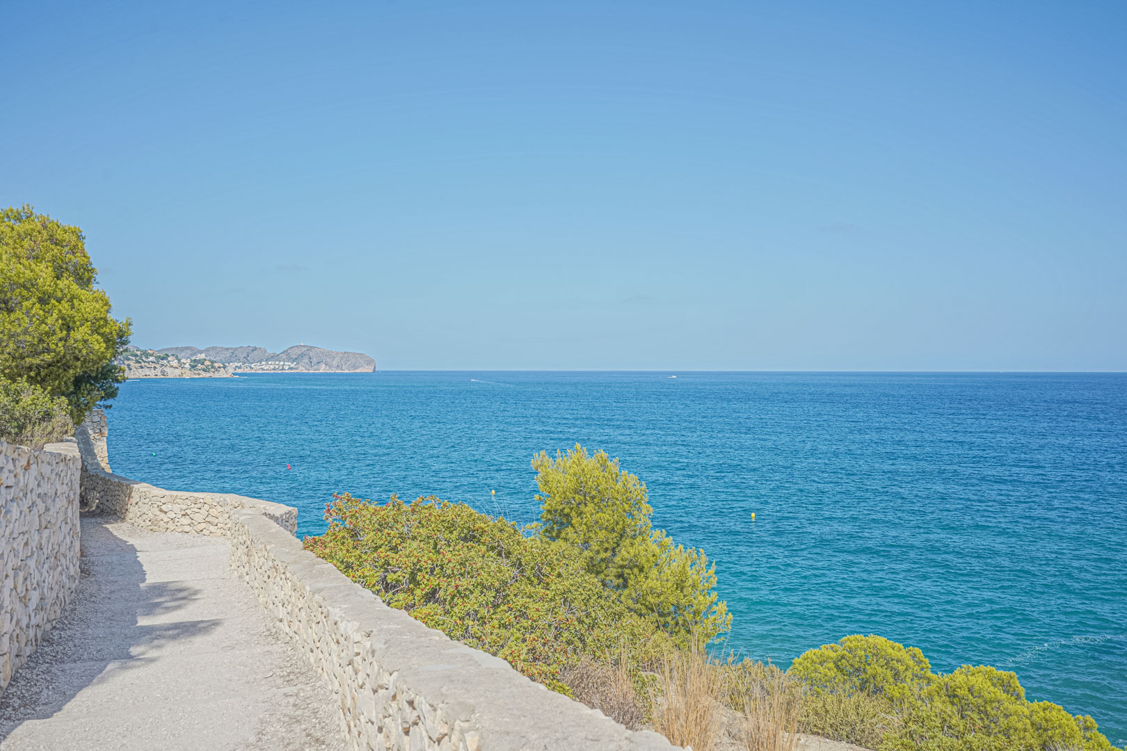 Coastal path featuring traditional dry stone walls and panoramic views of the blue Mediterranean Sea, bordered by native pine trees and shrubs.