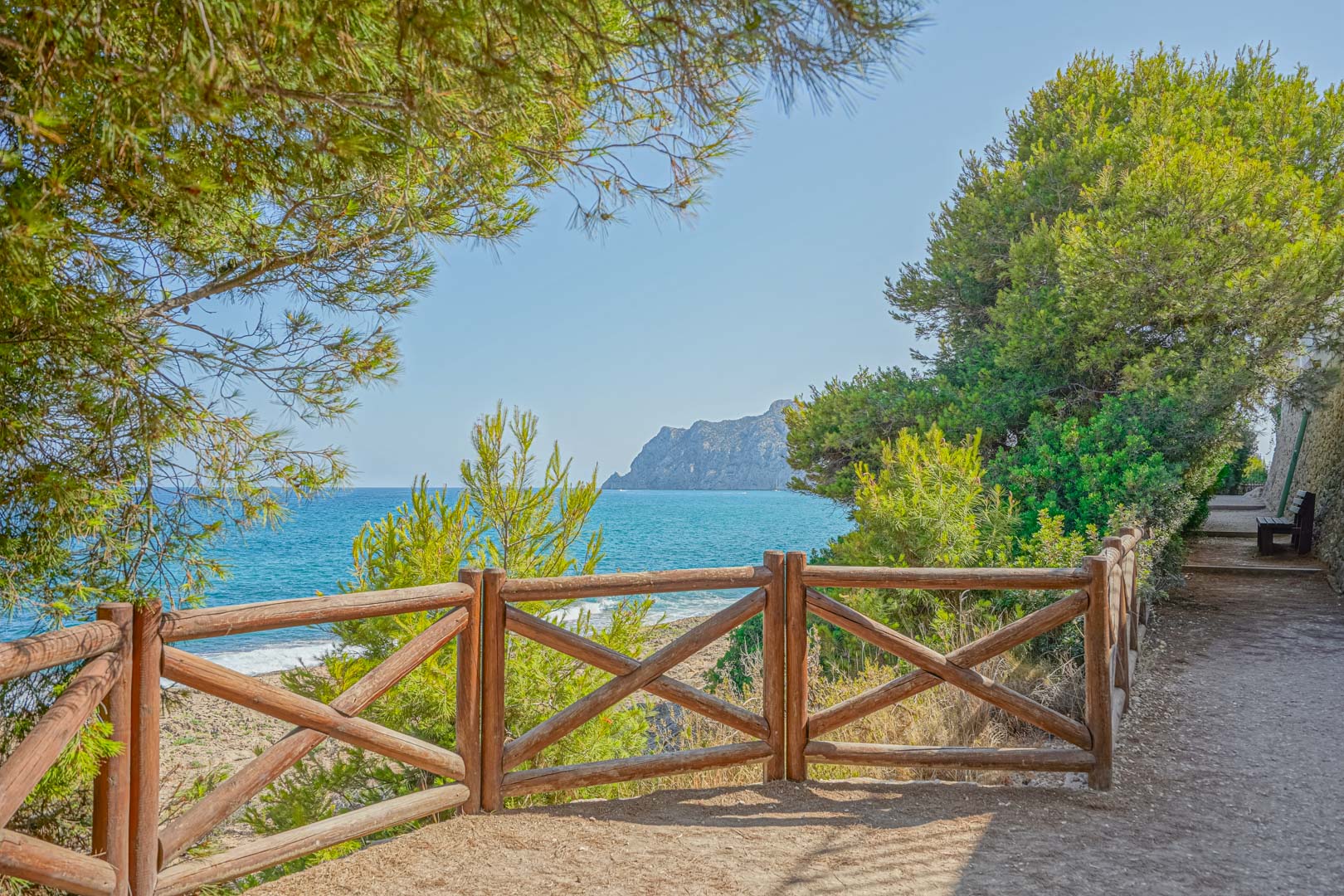 Coastal walking path featuring a rustic wooden railing and panoramic views of the Mediterranean Sea and the Peñón de Ifach framed by pine trees.