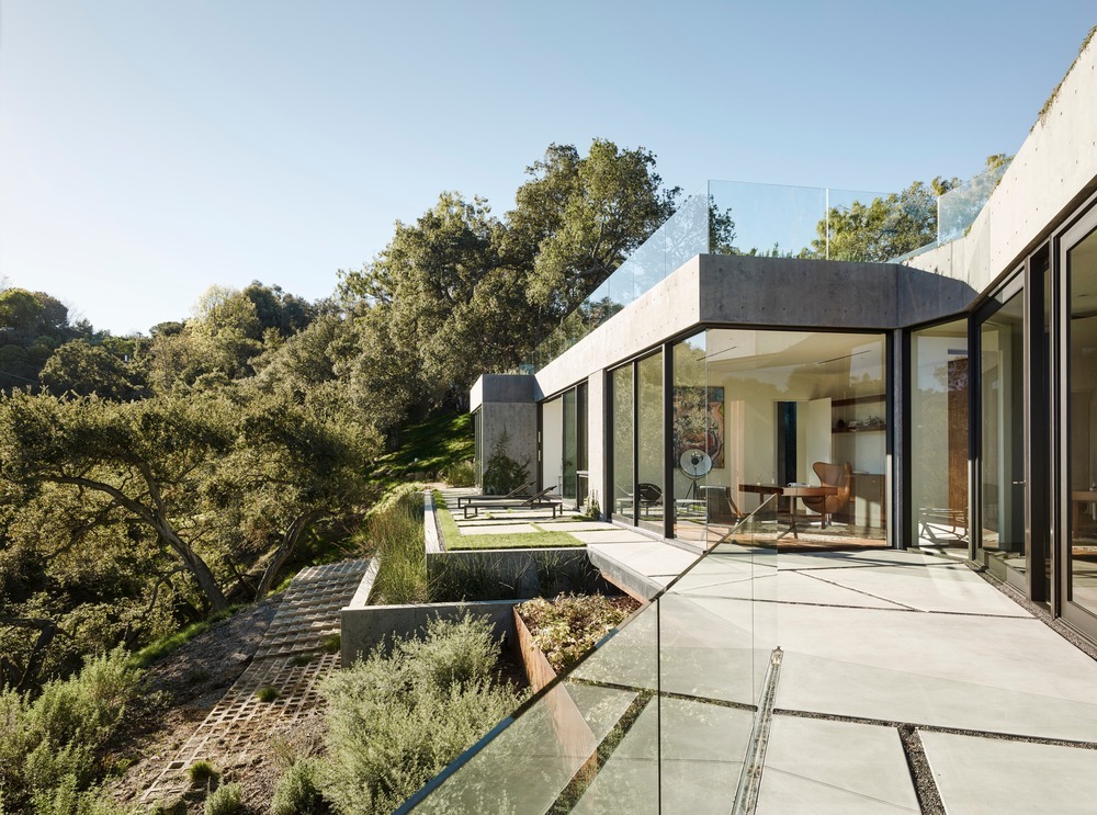 Modern concrete villa featuring floor-to-ceiling glass walls, a geometric paved terrace with glass railings, and views of the surrounding Mediterranean forest.