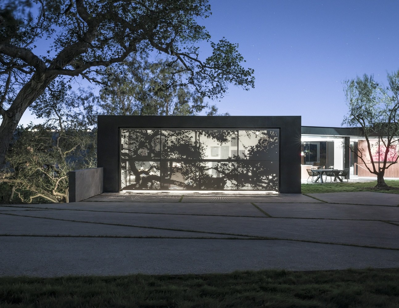 Modern facade featuring a backlit perforated metal screen with tree motifs, set against minimalist concrete paving and an integrated outdoor dining area at dusk.