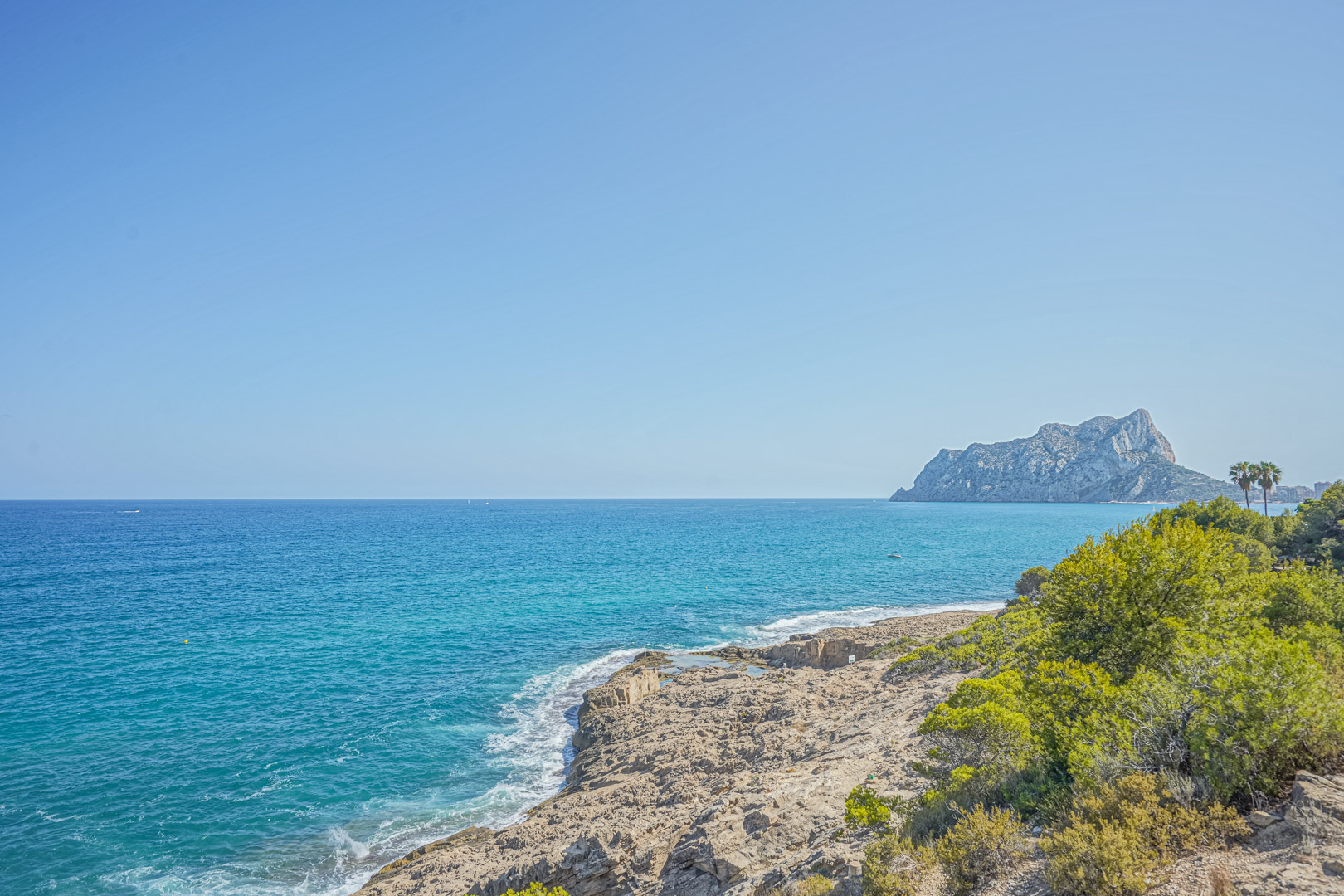 Panoramic view of the Benissa coastline featuring the Peñón de Ifach in the distance, clear turquoise waters, and rugged rocky cliffs with native pine vegetation.