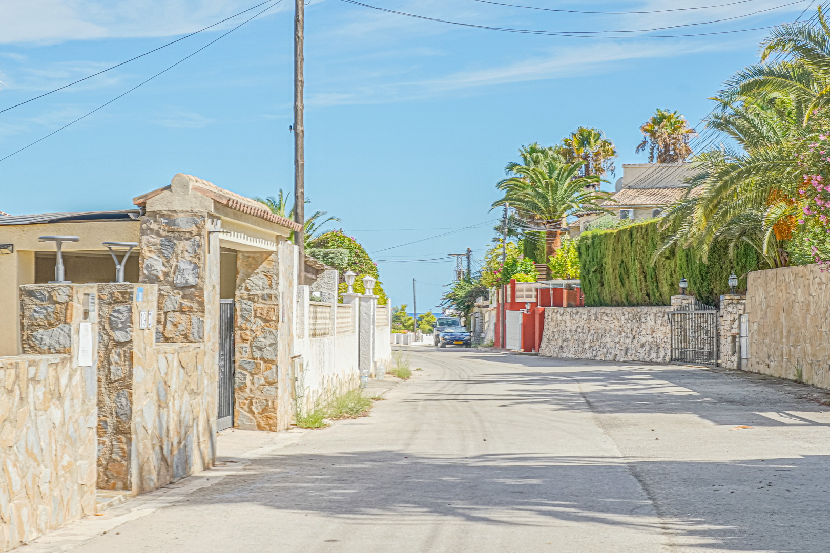 View of the residential access road lined with natural stone walls and Mediterranean greenery, offering a glimpse of the sea in the distance.
