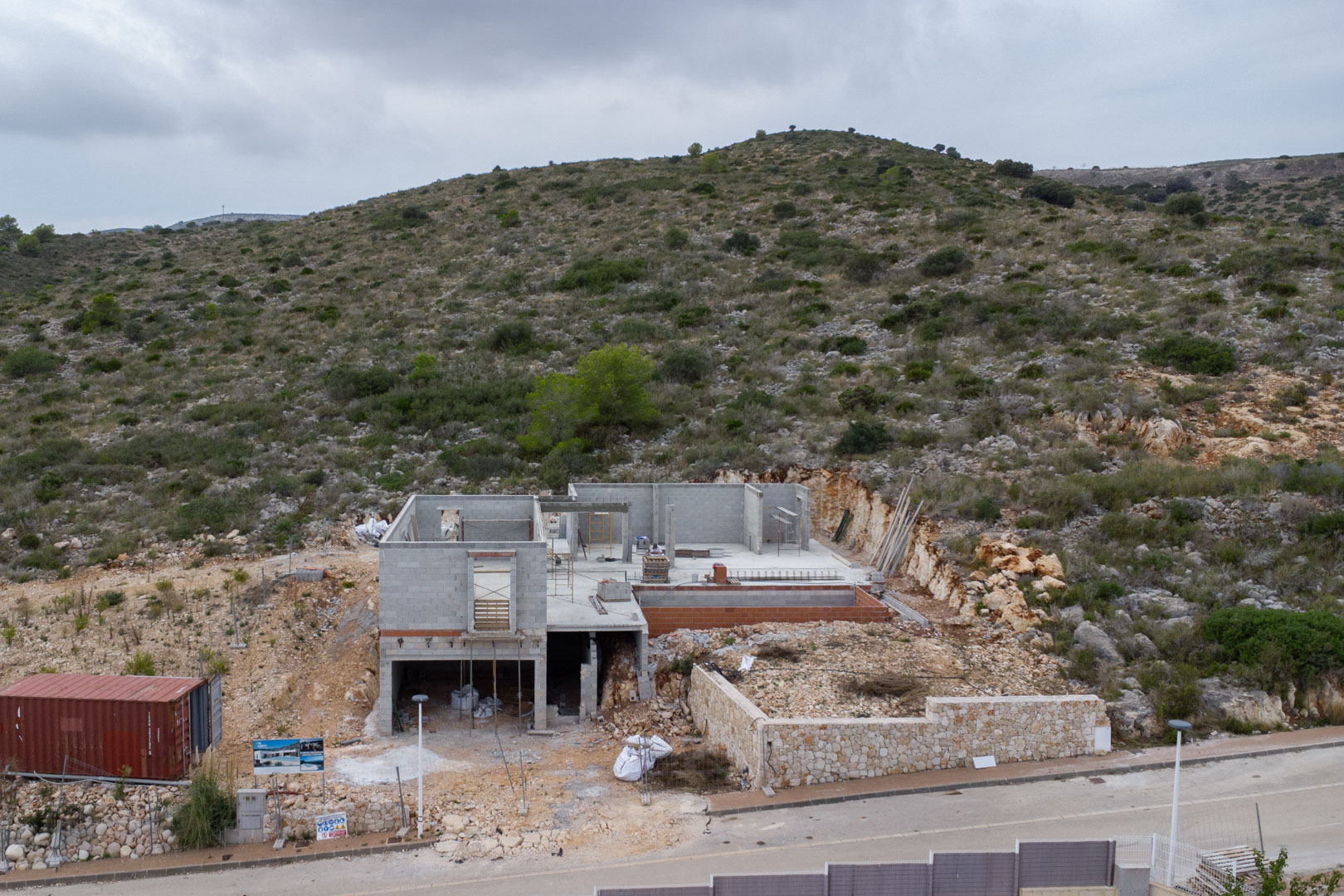 Construction phase of a multi-level villa in Pedreguer, featuring concrete block structures, a natural stone perimeter wall, and integration into the rocky hillside.