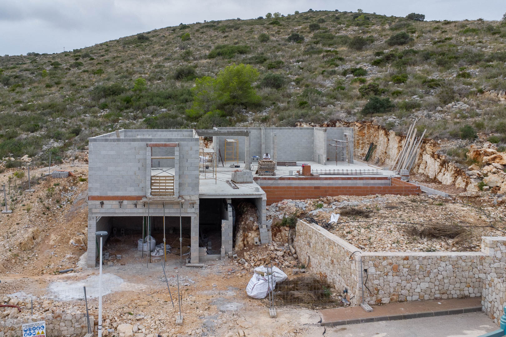 Current construction phase of a modern villa in Pedreguer, featuring concrete blockwork, stone retaining walls, and a hillside architectural layout.