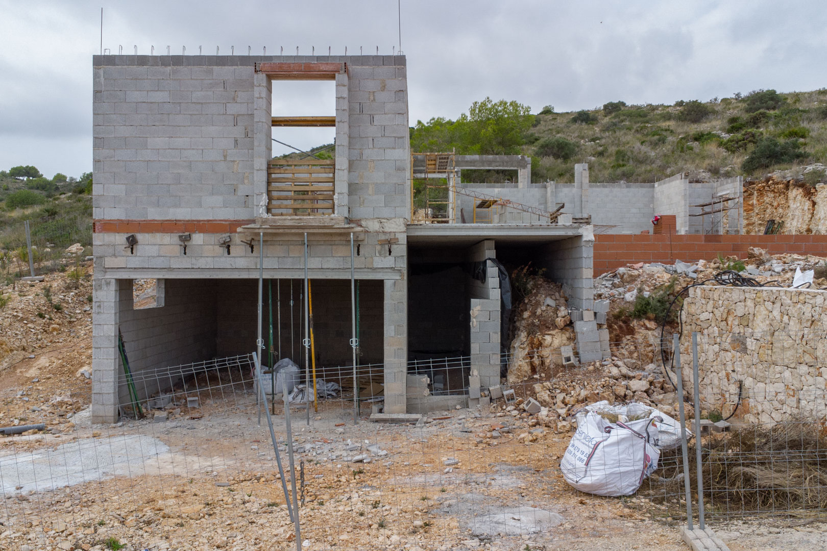Exterior view of the concrete block structure under construction, featuring multiple levels and a natural stone retaining wall on a hillside in Pedreguer.