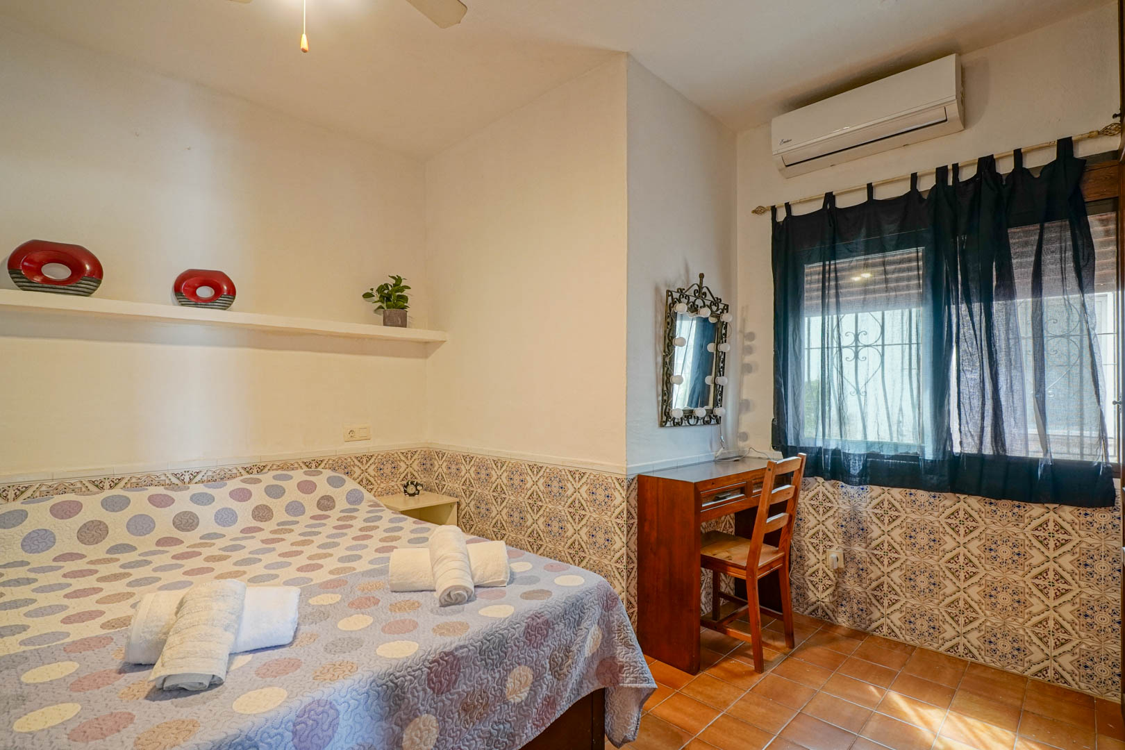 Bedroom featuring decorative wall tiling, a wooden vanity desk with a lighted mirror, and air conditioning. Finished with rustic terracotta floor tiles.